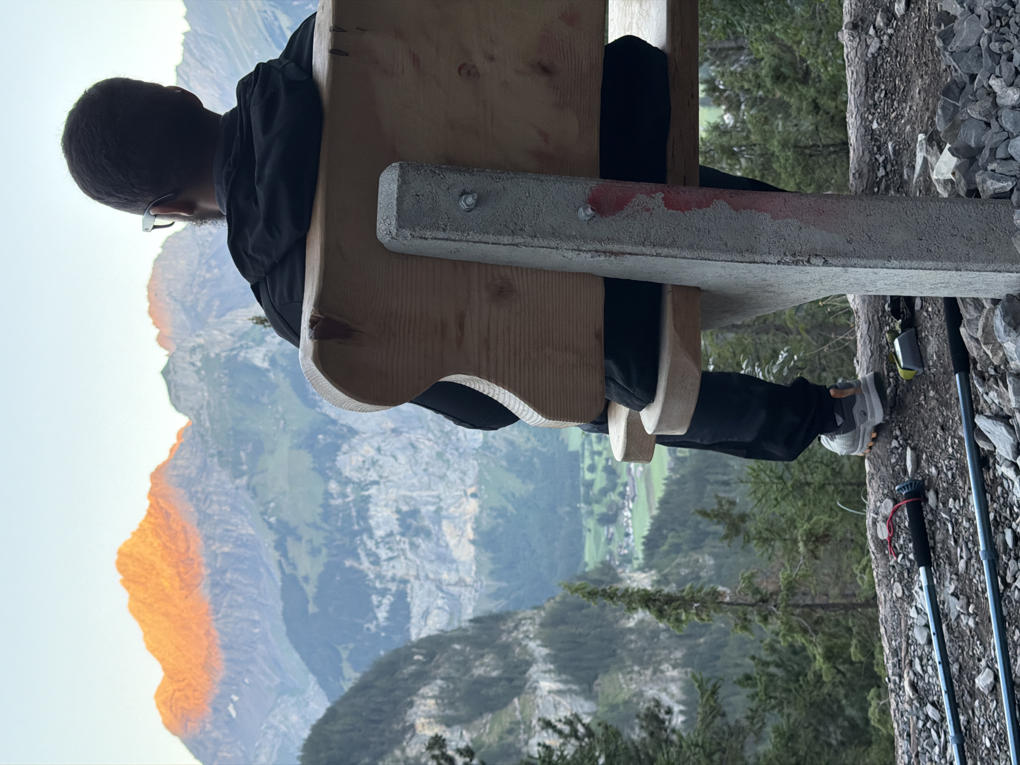 A person wearing glasses and a black jacket sits on a wooden bench with a concrete support, looking out at a mountain vista in Kandersteg, Switzerland. The sun is setting, casting an orange glow on the mountain peaks. In the foreground, hiking poles and rocks are visible. The environment is mountainous, with lush green slopes and trees.
