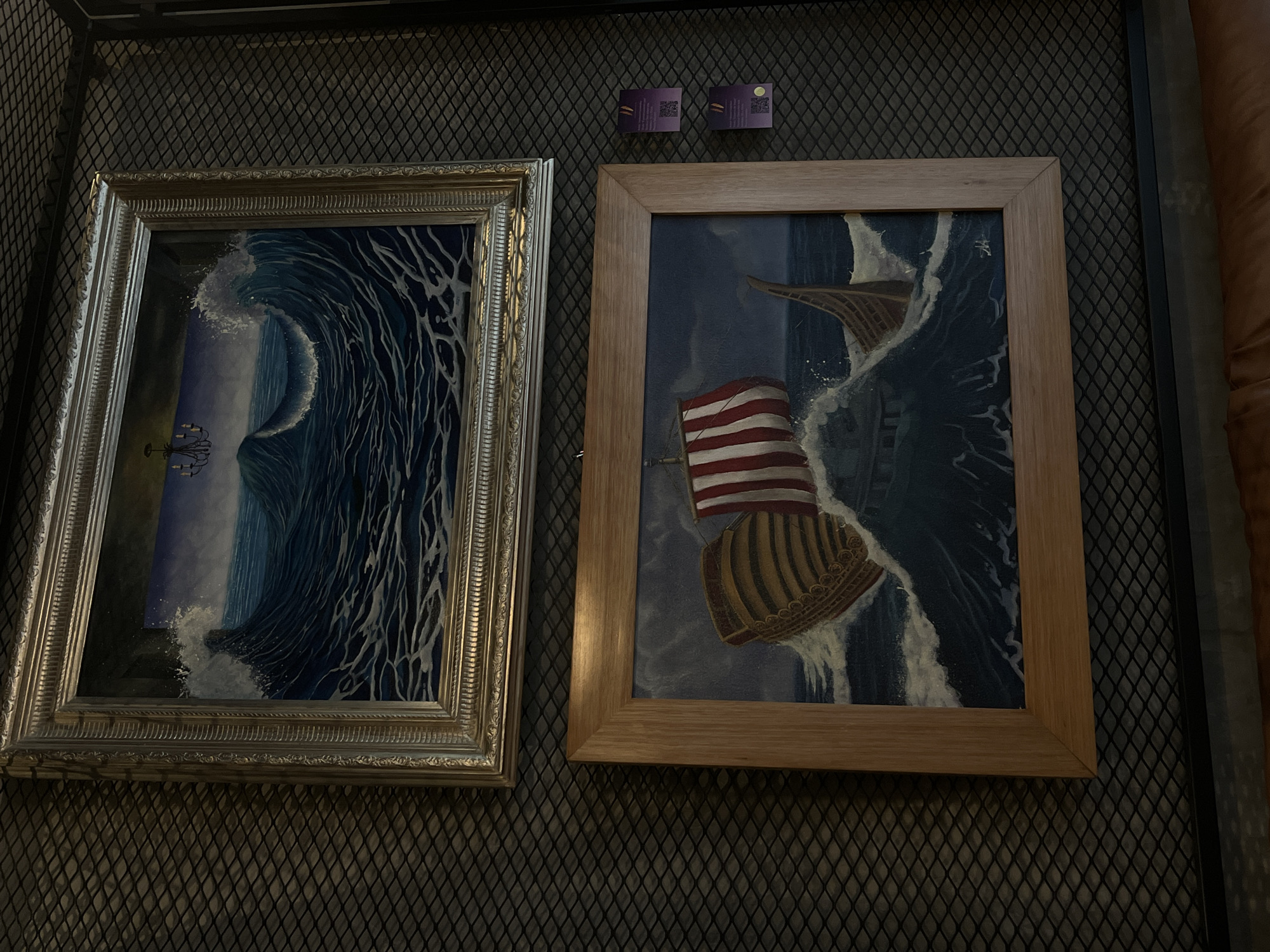 Two framed paintings hang on a dark, patterned wall. The painting on the left, framed in silver, depicts a large, curling wave near a shore where a chandelier is visible. The painting on the right, framed in light wood, features a ship caught in turbulent waves with a prominent American flag. Above the paintings are two small, purple squares with QR codes or similar patterns. To the right of the second painting, a textured brown surface is visible, possibly furniture or another wall. The location is Unknown, Unknown.