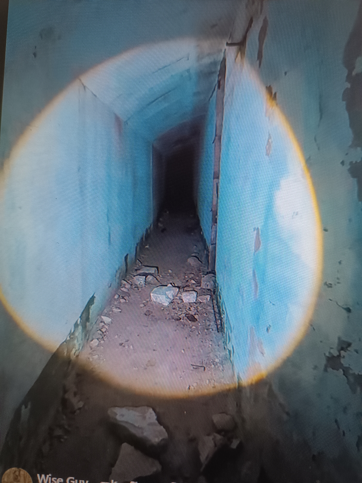 The image shows a narrow, dimly lit concrete tunnel with a rough, debris-strewn floor. The walls are painted a faded blue and appear damaged, with peeling paint and exposed concrete. A bright, oval-shaped light source, possibly a flashlight, illuminates the foreground and the left wall of the tunnel, casting a warm glow that fades to a pale blue on the distant walls. The tunnel recedes into darkness at the far end. The bottom of the image shows what appears to be a digital interface with text that includes "Wise Guy". This scene is located in the Tbilisi metro system, likely an abandoned or disused section.