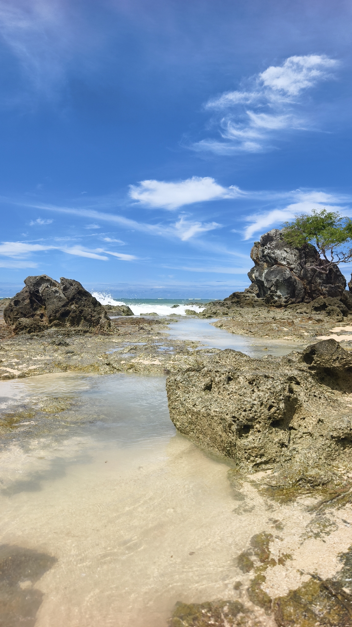 The image depicts a coastal scene in Kute, Indonesia. The foreground features a rocky shoreline with shallow pools of clear water over a sandy base. Various sizes of rocks are scattered throughout, some partially submerged. In the midground, ocean waves are visible, breaking on the rocks. Two prominent rock formations are present: one on the left and a larger one on the right, supporting a small tree. The background consists of a bright blue sky with wispy white clouds. There are no people visible in the scene.