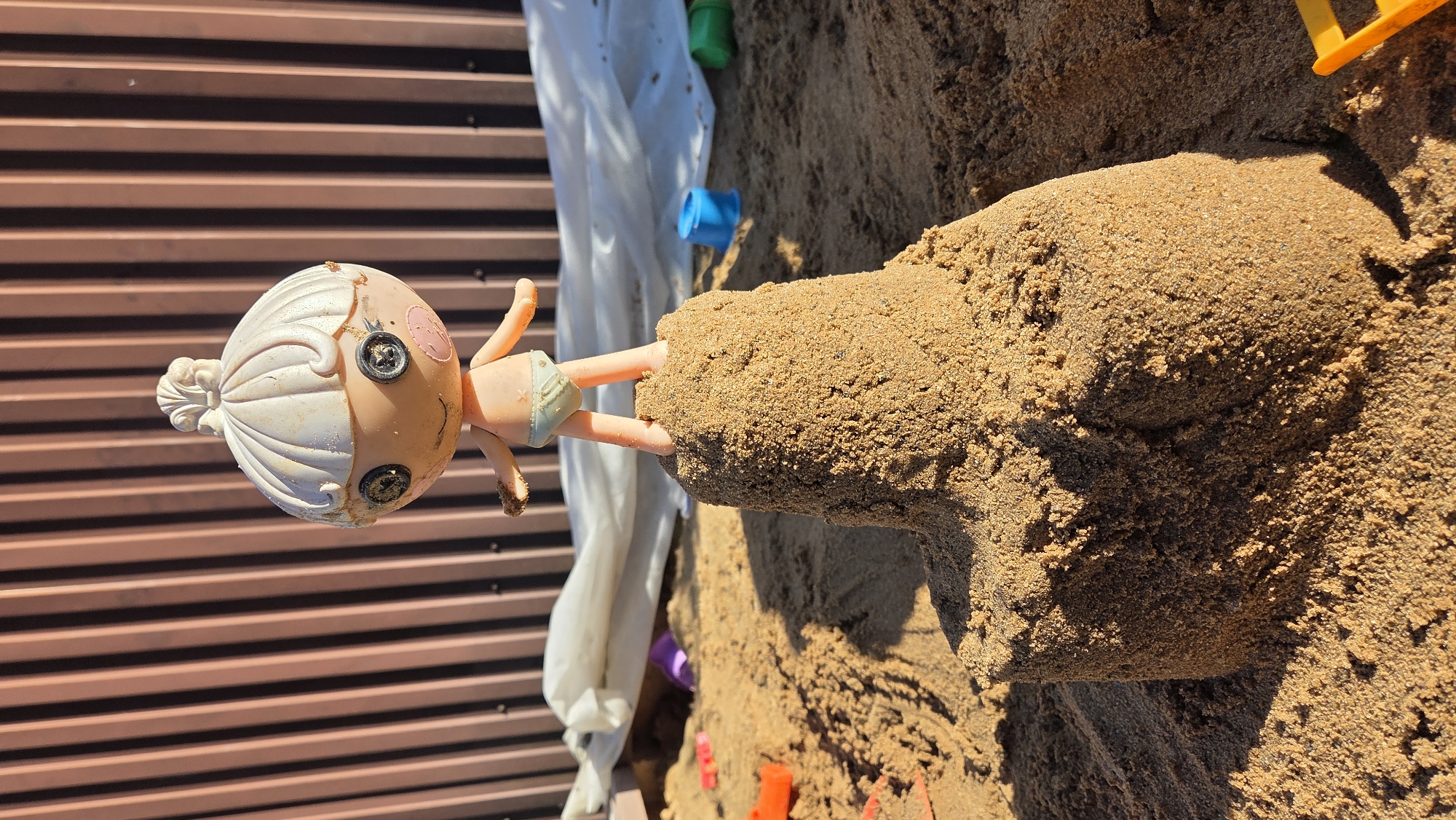 A doll stands atop a sandcastle, which is situated in a sandbox. The doll has a white topknot hairstyle and button eyes. It is wearing a light blue diaper. The sandcastle is brown and roughly conical in shape. Other toys, including a purple object, an orange toy, a blue bucket, and a green bucket, are scattered in the sand around the sandcastle. In the background, a brown corrugated fence is visible, with white fabric draped over a structure behind the sandcastle. The scene is outdoors, under direct sunlight. The location is specified as Razdol'e, Russia.