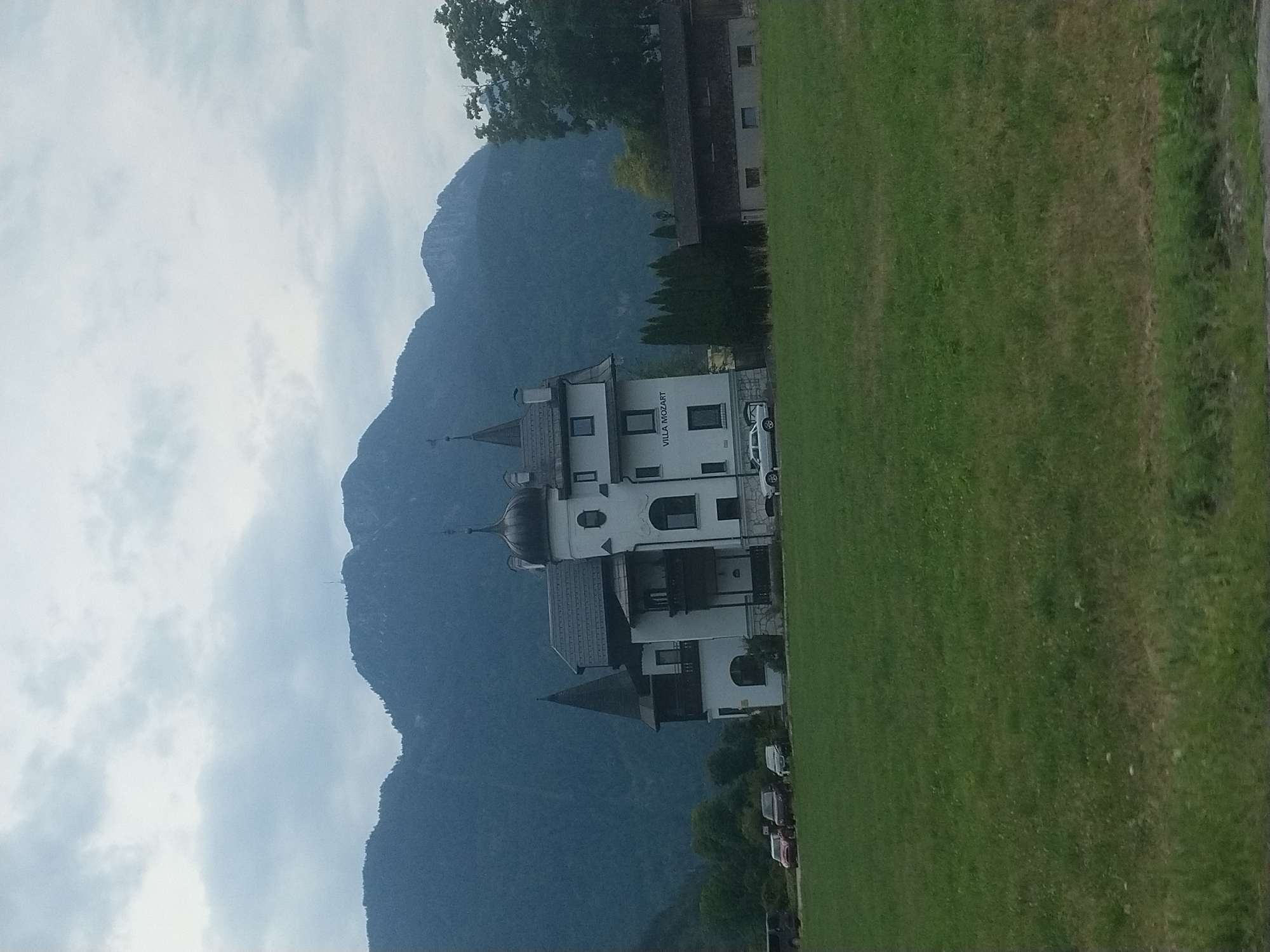 The image shows a grand white building with dark grey roofs, identified by the signage "VILLA MOZART" on its facade. This villa, located in Bad Ischl, Austria, is situated in front of a majestic mountain range under a cloudy sky. In the foreground, a lush green lawn stretches out, with a few cars parked in front of the villa and a smaller, dark-colored building to its right. The overall scene depicts a serene, picturesque setting that combines architectural grandeur with natural beauty.