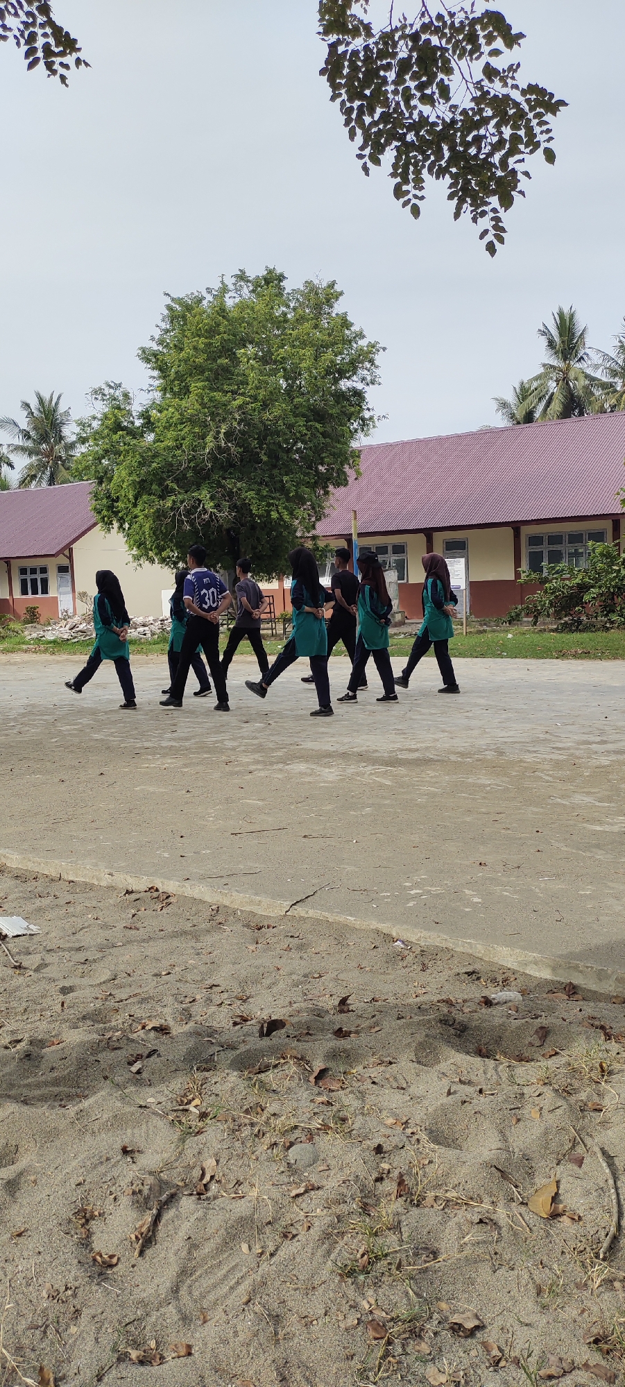 An outdoor, eye-level shot captures a group of eight young people walking in single file across a sandy and concrete outdoor area in front of a school building. The individuals, some wearing hijabs, are dressed in matching green tops and dark pants. The school building features a red-tiled roof and yellow and brown walls with windows. Trees, including a large one behind the group and palm trees on either side of the building, dot the landscape. The sky is overcast.