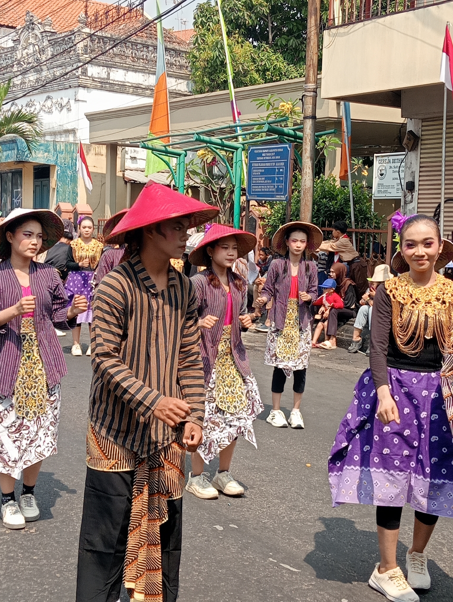 In the city of Bintoro, Indonesia, a group of people are participating in a parade. The central focus of the image is a young man and several young women dressed in traditional attire. The man, in the foreground, wears a black outfit with thin brown pinstripes, a red conical hat, and a patterned sarong. The women are adorned in colorful, patterned clothing, including long skirts and jackets, and wear straw hats. One woman on the right is particularly striking with elaborate gold jewelry around her neck and a bright purple skirt. The parade appears to be taking place on a paved street lined with buildings and trees. In the background, flags are visible, including the red and white Indonesian flag. Spectators are present on the sidewalks and in the background, some watching the parade, others casually conversing. The atmosphere seems festive and celebratory, indicative of a cultural event or festival in the city.