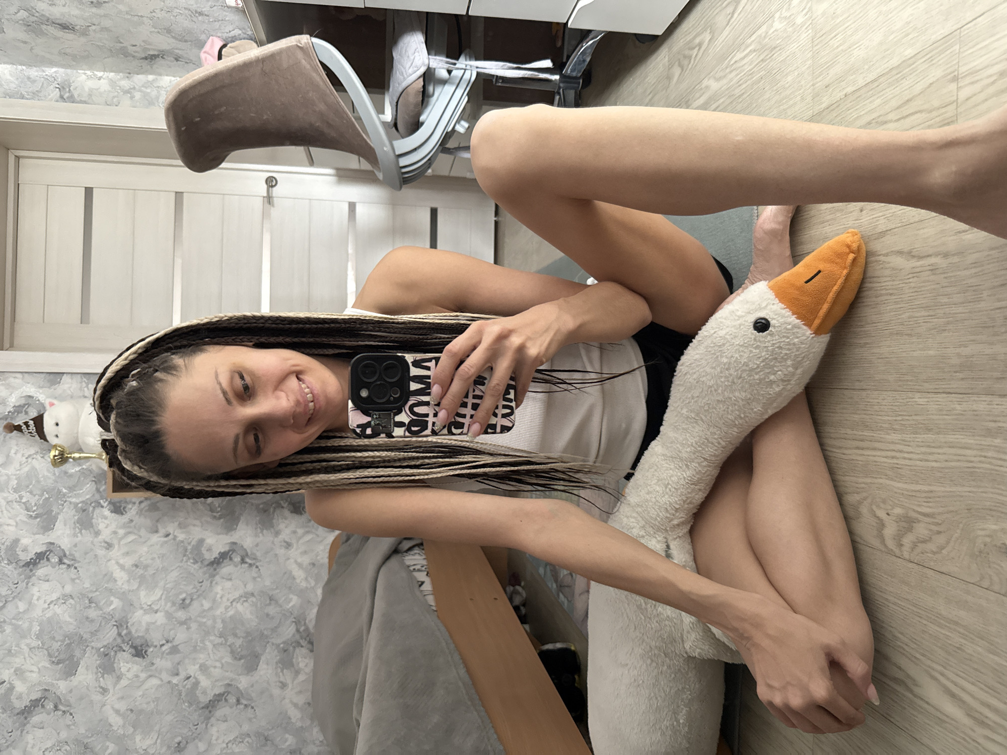 A young woman is depicted indoors, seated on a light wooden floor in what appears to be a casual residential setting in Rusanovo, Russia. She has long, multi-toned braids, a mix of blonde and dark strands, and is wearing a white tank top and dark shorts. She holds a black smartphone in her left hand, taking a selfie, and smiles cheerfully towards the camera. A large, fluffy white plush toy, shaped like a goose with an orange beak and black eyes, is nestled between her legs and partially embraced by her right arm.
The background features a textured grey and white wall to her left, and a white paneled door with a silver handle to her right. Visible in the upper background are parts of a light brown chair with a fabric cover and a white desk or cabinet. A small white snowman figurine with a brown hat is perched on a shelf behind her head. The lighting suggests it is daytime. Her phone case is black with white text, partially visible, which appears to read "YOU ARE MINE". The woman has light-colored nail polish on her fingers. The overall scene conveys a relaxed and happy atmosphere.
