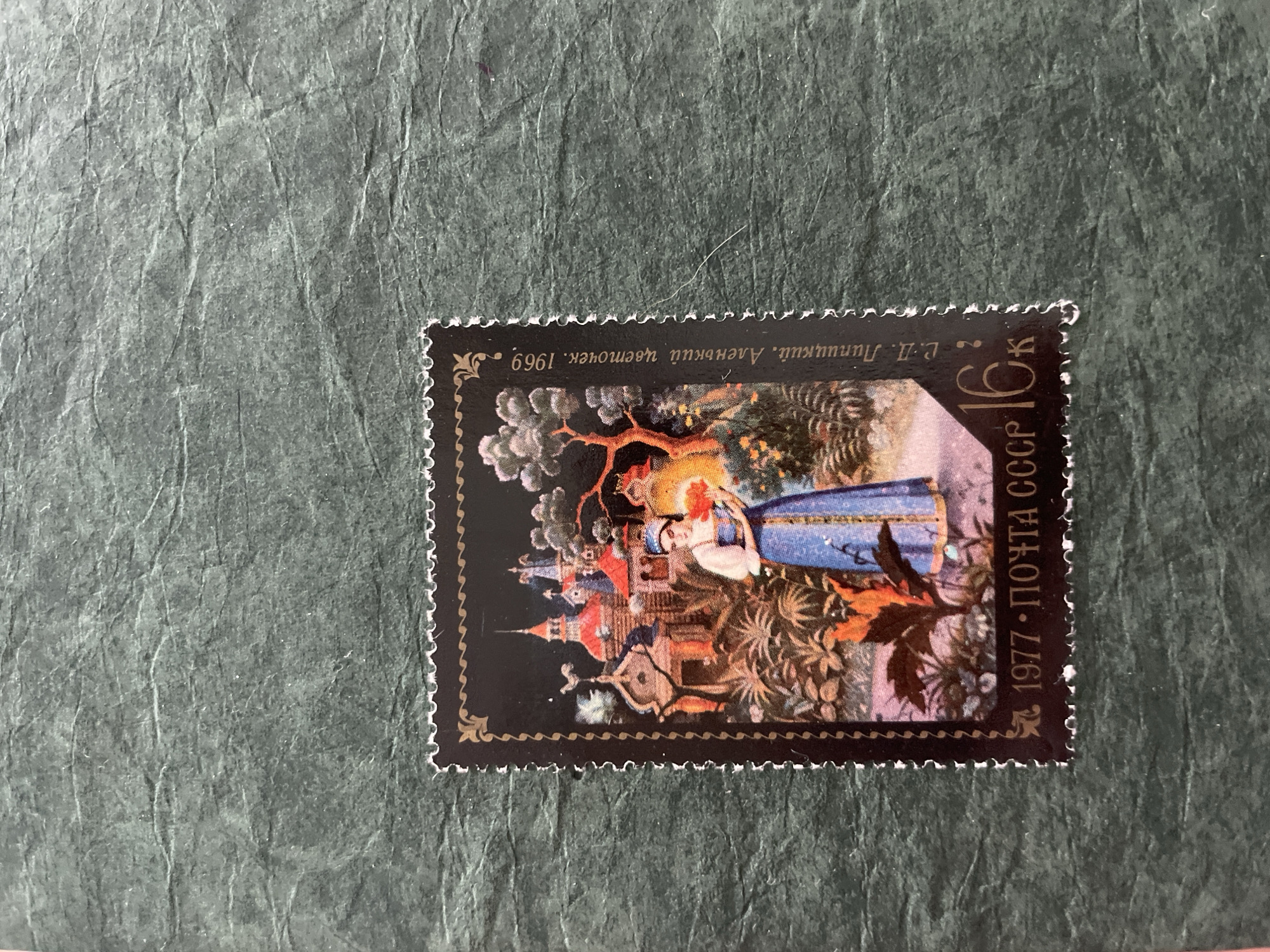 The image is a close-up of a postage stamp on a dark green, textured background. The stamp features a colorful illustration with a dark background. In the foreground, a woman in a blue traditional dress and white head covering stands holding a bouquet of red flowers. She is positioned in front of a lush garden with various plants and foliage. Behind her, a whimsical architectural structure, resembling a castle or temple with red roofs and spires, is depicted. The sky is dark, suggesting either night or a stylized depiction. Along the right edge of the stamp, text in Cyrillic script reads "Е.Л. Липицкий, Аленький цветок. 1969". Along the bottom edge of the stamp, the year "1977" and "ПОЧТА СССР" are visible, along with a value of "16 К". The background appears to be a textured paper, possibly a scrapbook page or album. There is no visible depiction of the city of Sokol, Russia within the stamp's illustration.