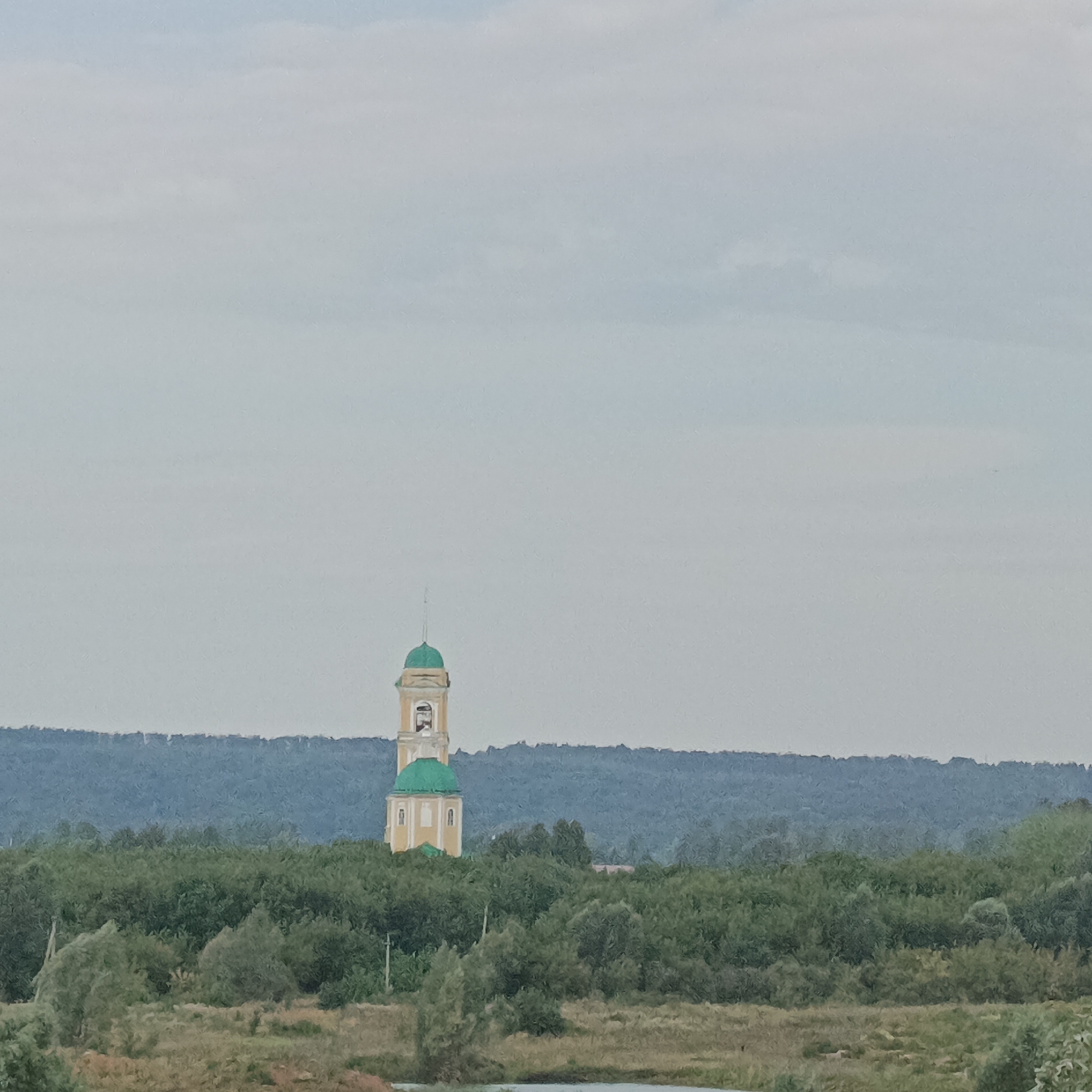 A bell tower with green domes stands tall amidst lush green trees, silhouetted against a pale, overcast sky. The tower is light-colored, possibly yellow or beige, with architectural details that suggest a religious structure. In the foreground, a field of dry grass stretches towards a body of water, hinting at a rural or semi-rural landscape. The backdrop features a dark blue, hazy mountain range. The scene is peaceful and expansive, typical of the Russian countryside, potentially the village of Nikolo-Beryozovka, which is known for its natural beauty and religious sites.