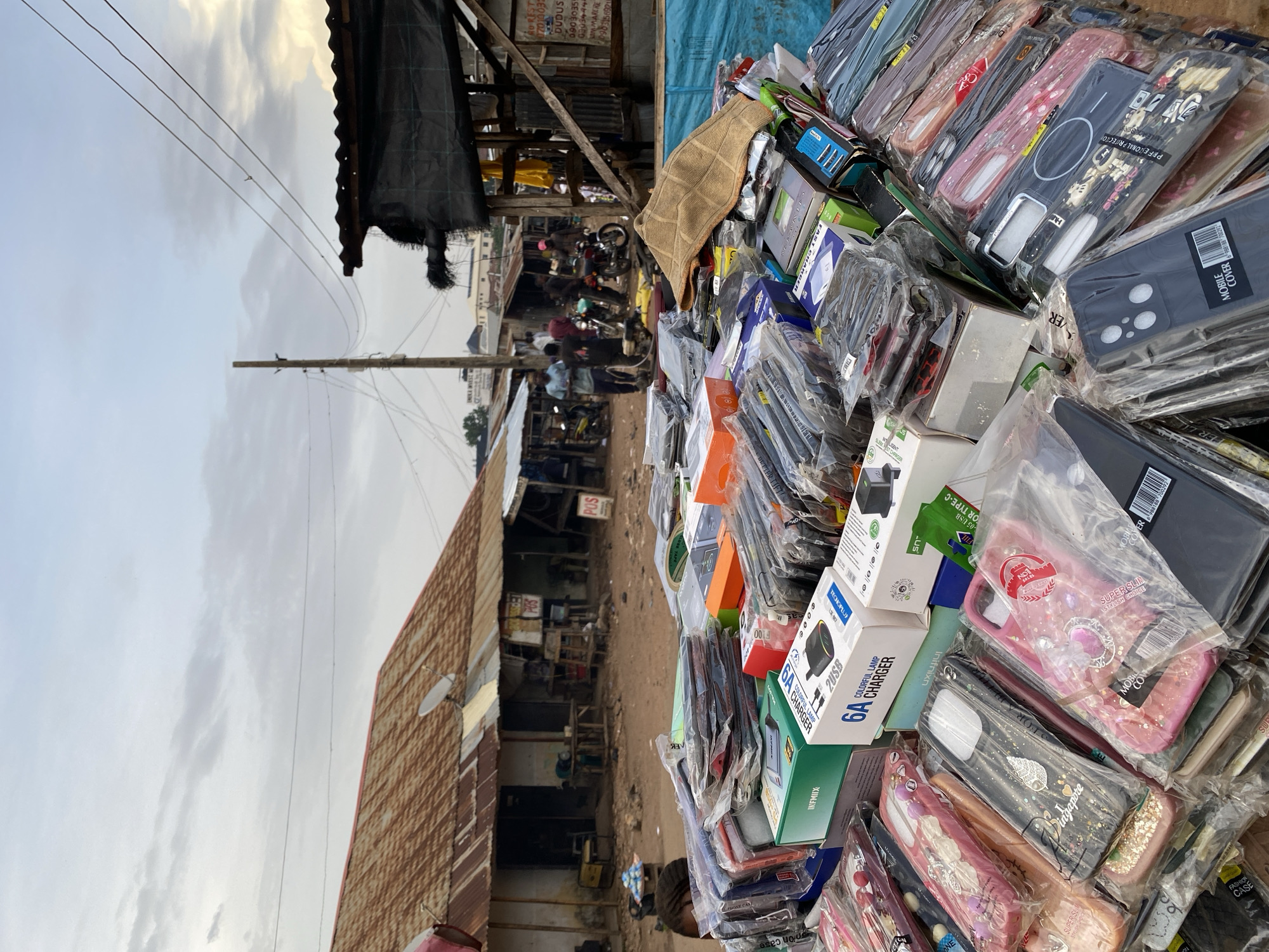 The image depicts an outdoor market scene in Bukuru, Nigeria. In the foreground, a large assortment of mobile phone accessories, primarily phone cases and chargers, are piled high and displayed for sale. These items are individually packaged in clear plastic. Among the accessories, several boxes are visible, including "INFINIX" and "6A COLORFUL LAMP CHARGER 2USB". In the mid-ground, there is a dirt path leading to a row of market stalls with corrugated metal roofs. Some stalls appear to be simple structures with open fronts, showcasing goods within. A few people are visible in the background, some appearing to be vendors or shoppers, and motorcycles are parked near the stalls. Overhead, power lines crisscross the sky, and the sky itself is partly cloudy with a pale, overcast light, suggesting it might be late afternoon or early morning. The overall impression is of a bustling, informal marketplace.