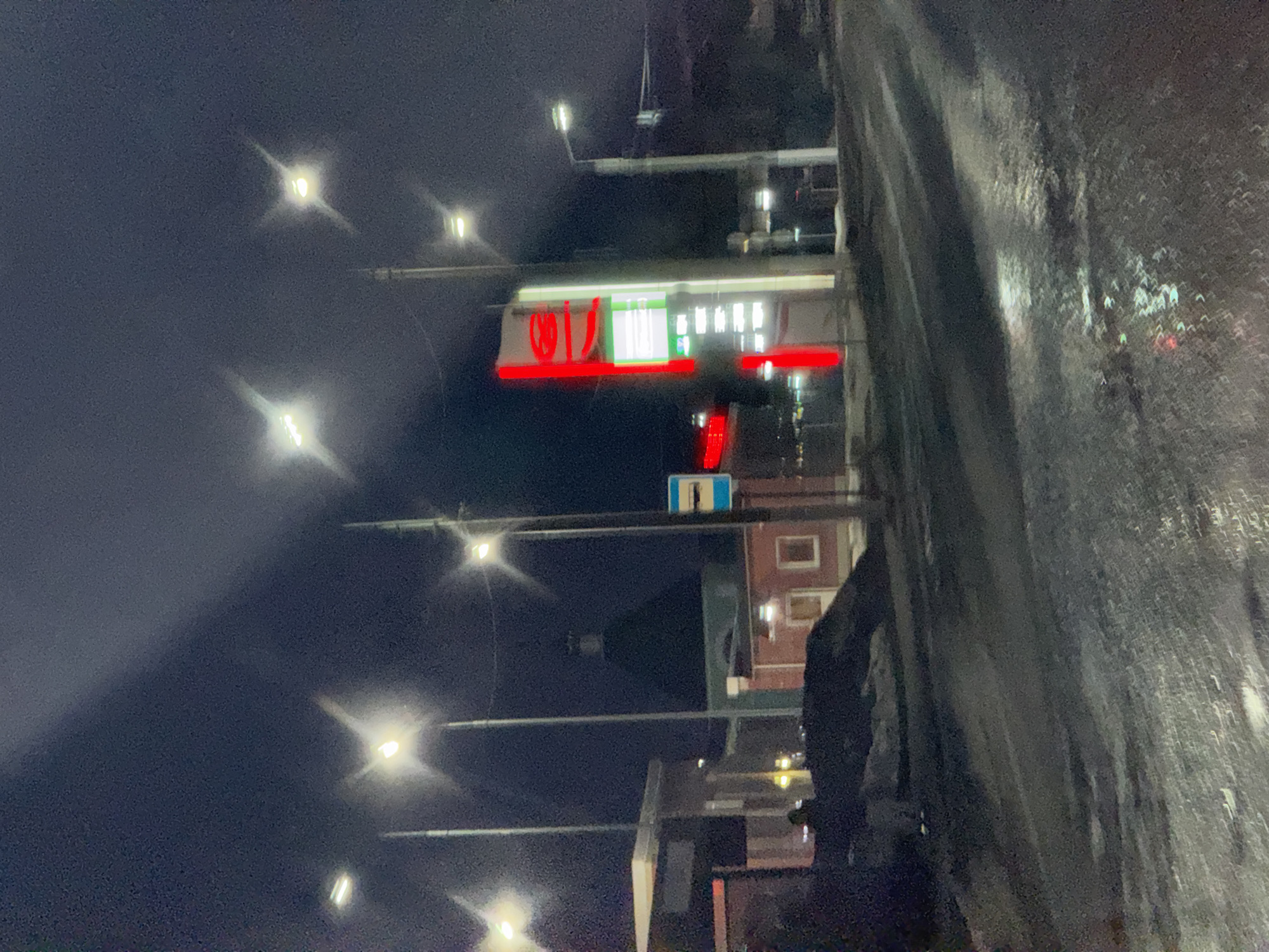 The image shows a street in Chaikovsky, Russia, at night. Streetlights line the road, illuminating a wet asphalt surface. Prominently visible is a brightly lit convenience store with a red and white sign. The architecture of the store suggests a modern design. A road sign is also present. The sky is dark and overcast. The overall environment appears to be urban, with buildings visible in the background.