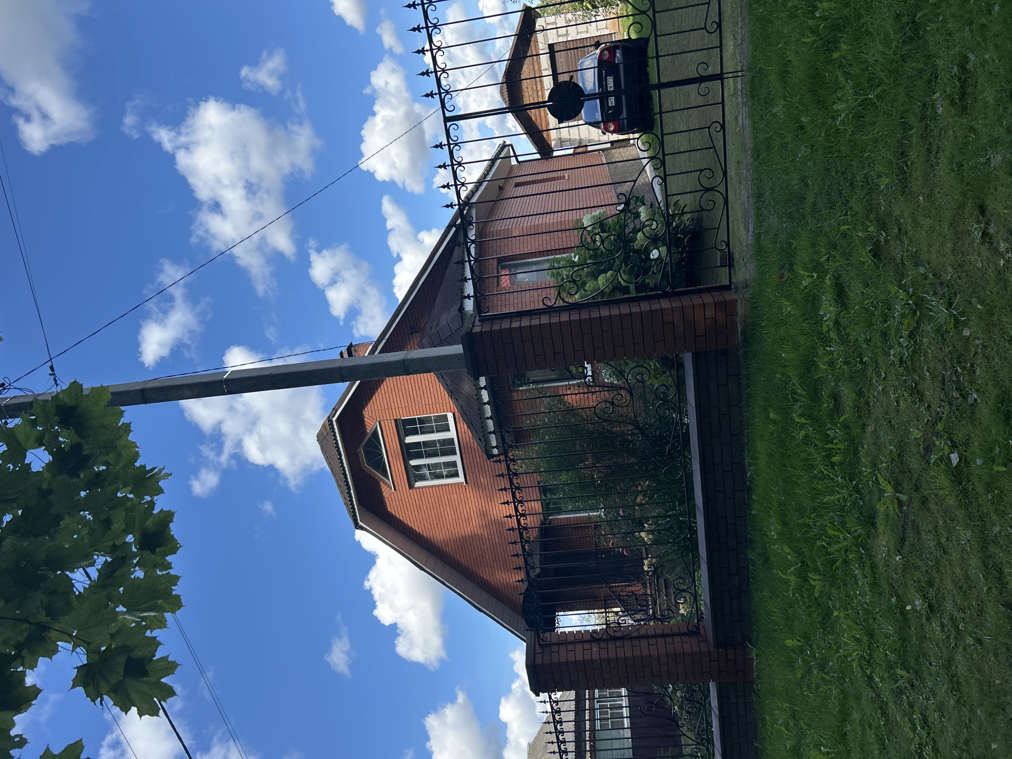 A red brick house with a dark roof is visible in the city of Kalitsino, Russia. The house has a dormer window with multiple panes and a triangular decorative window above it. A black metal fence with decorative scrollwork surrounds the property. In front of the house, there is green grass and some flowering bushes. A dark-colored car is parked in front of a detached garage made of light-colored bricks and wood. A black utility pole stands to the right of the house, with power lines running overhead. The sky is blue with scattered white clouds. The image appears to be taken on a sunny day.