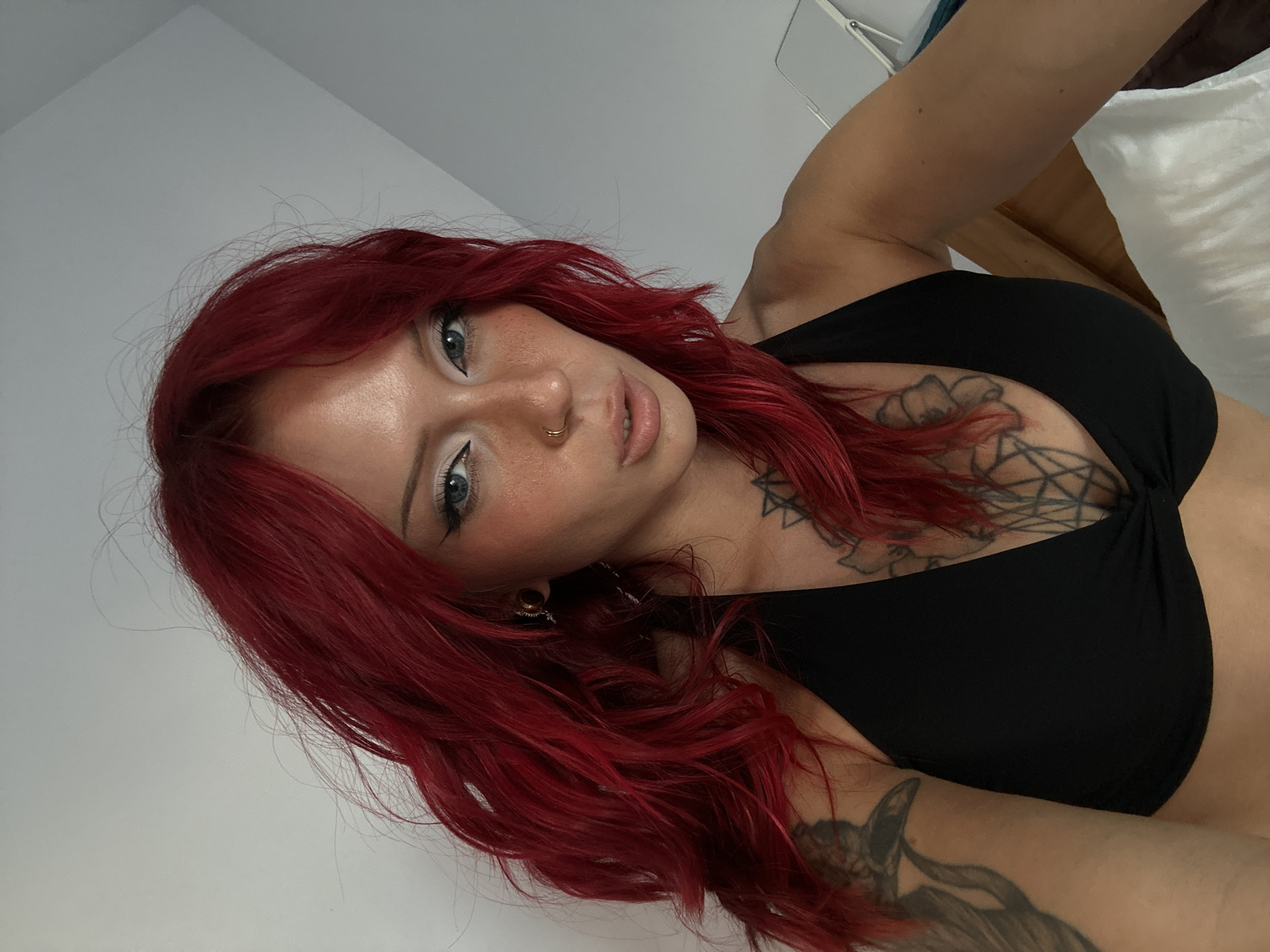 A woman with bright red hair and a black top is looking at the camera in what appears to be a bedroom. She has a nose ring and tattoos on her chest and arm. The background is a plain white wall. The scene does not provide enough information to determine the location in North Kuta, Indonesia.