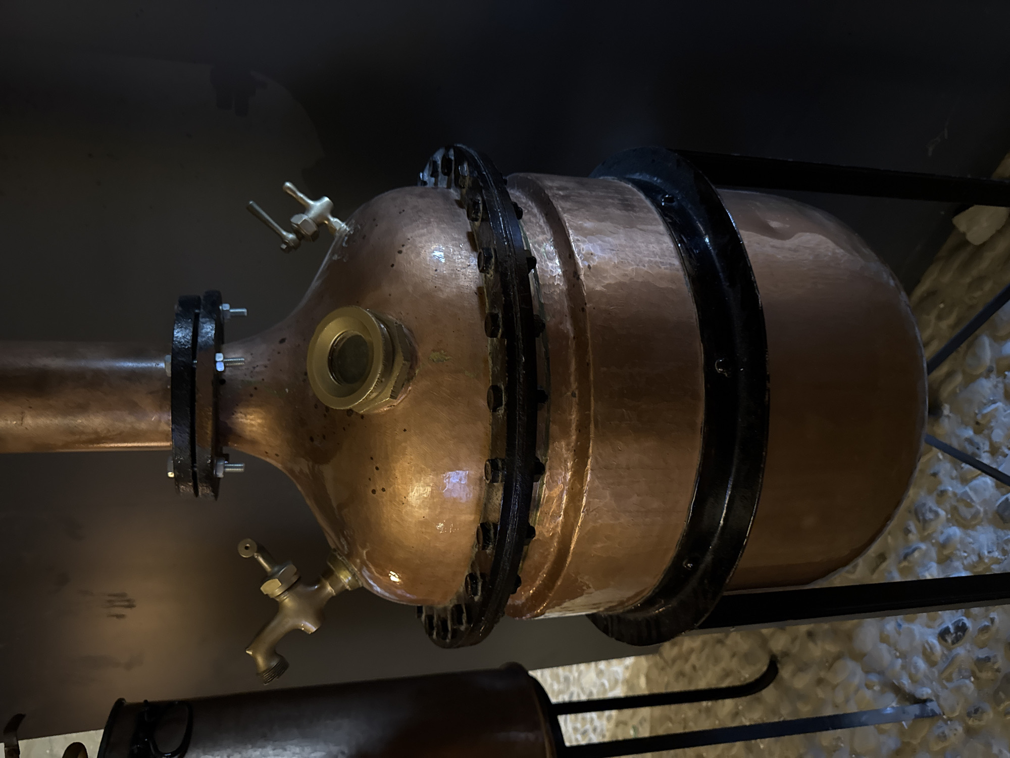The image shows a close-up, slightly elevated, eye-level view of a copper alembic, a piece of distillation equipment, likely used for producing spirits or essential oils. The alembic is made of polished copper and features a bulbous lower section connected to a cylindrical upper section via a flanged joint secured with bolts. Two brass spigots or valves are visible on the upper bulbous part, one on the left and one on the right. A round sight glass with a brass housing is also present on the upper bulbous section, positioned centrally. The alembic stands on a black metal frame. To the left, a portion of another dark metal cylindrical object is visible, also on a dark metal stand, with a handle. The background is a dark, nondescript wall, and the floor appears to be covered in light-colored, uneven stones or pebbles. The lighting is dim, highlighting the metallic sheen of the copper. The scene is indoors, suggesting a workshop, distillery, or a display in a historical or museum setting. The location is identified as Dolceacqua, Italy.