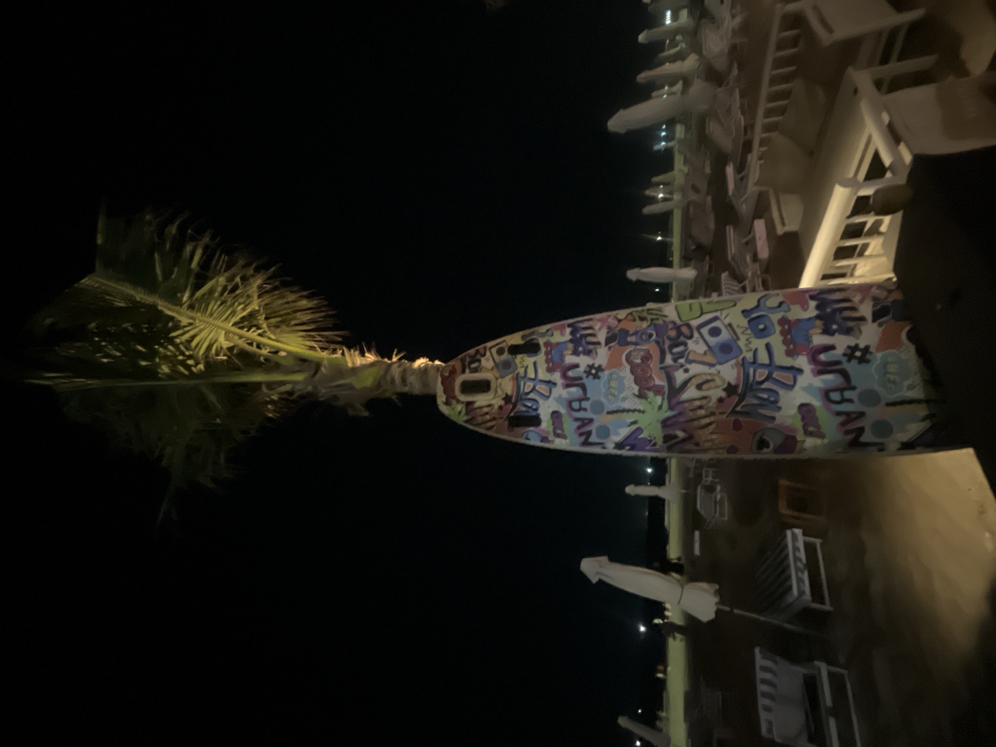 This image captures a scene at night, likely on a beach or waterfront area in Da Nang, Vietnam. The primary subject is a palm tree, its fronds illuminated and appearing somewhat windswept against a dark sky. To the right of the palm tree, a large, cylindrical object is visible, covered in a vibrant, graffiti-style art with various words and symbols. Some of the visible text includes "URBAN," "#," and what appears to be "ROCK."
Beyond the palm tree and the decorated object, rows of empty white lounge chairs and umbrellas suggest a public space that is closed for the night. These chairs are neatly arranged, hinting at a resort, hotel, or beachside establishment. The lighting in the scene is artificial, casting a yellowish glow on the chairs and the ground, and creating reflections that suggest the presence of water or a wet surface nearby. Small, distant lights are visible in the background, potentially indicating buildings or a cityscape.
The overall mood is quiet and still, as expected for a nighttime scene with no people present. The dark sky and artificial lighting suggest it is after sunset, and the absence of any visible weather phenomena implies clear conditions. The presence of palm trees and lounge chairs strongly points to a tropical or subtropical coastal location.