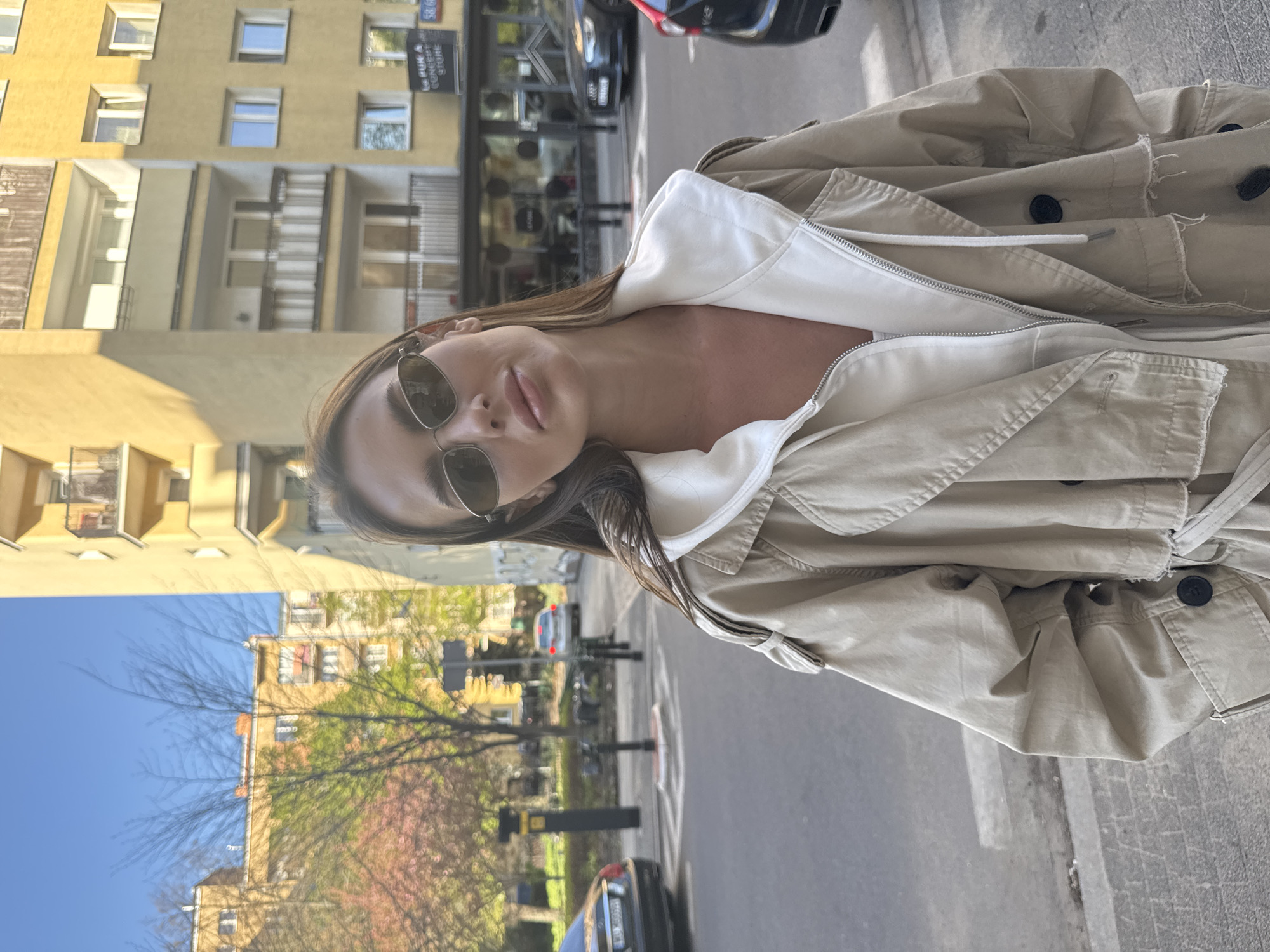A young woman is captured in a headshot-style image, likely a selfie, taken outdoors on a sunny day in an urban setting. She is positioned slightly to the right of the frame, looking directly at the camera with a neutral, posed expression. Her long brown hair frames her face, and she wears dark sunglasses. She is dressed in a light beige or khaki trench coat, worn open, revealing a white zippered hoodie or top underneath. A noticeable reddish mark is present on her upper chest and neck, potentially from sun exposure.
The background shows a bustling city street. To the left, a multi-story building with a warm yellow facade features numerous windows and balconies. To the right and further back, a paved street is visible with parked cars, including a dark sedan in the foreground and a silver car further down. Trees with green and pinkish foliage line the street, suggesting spring. A clear blue sky is visible in the lower left portion of the image. On a building in the background, a sign reads "PUKA CONCEPT STORE" and "58 56". The bright lighting indicates it is daytime, likely late morning or afternoon. The scene depicts a casual moment in a city environment, consistent with Warsaw, Poland.