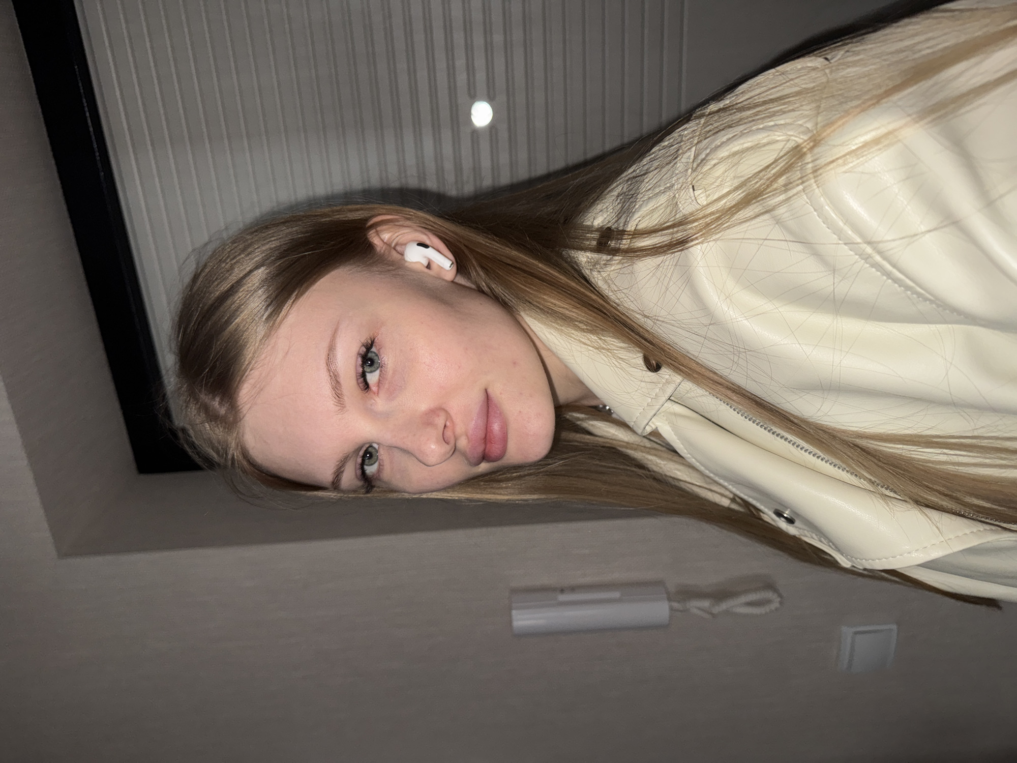 A portrait-style image, likely taken from below, features a young woman with light brown hair and light-colored eyes, possibly blue or green. She has fair skin and is wearing a cream or off-white leather or faux-leather jacket, parts of which show quilted stitching and a zipper. A white wireless earbud (AirPod) is visible in her left ear. She looks directly at the camera with a neutral, calm expression, her head slightly tilted.
The background consists of an indoor setting with a light-colored ceiling or upper wall, which has faint vertical ridging or paneling. A small, circular light source or fixture is visible on the ceiling, glowing faintly. To the lower right, a white rectangular intercom unit or thermostat is mounted on the wall, with a coiled wire extending from it. A dark vertical strip, possibly part of a doorframe or architectural element, is visible on the far left. The overall impression is an intimate, candid moment indoors in Perm, Russia. No visible text is present.