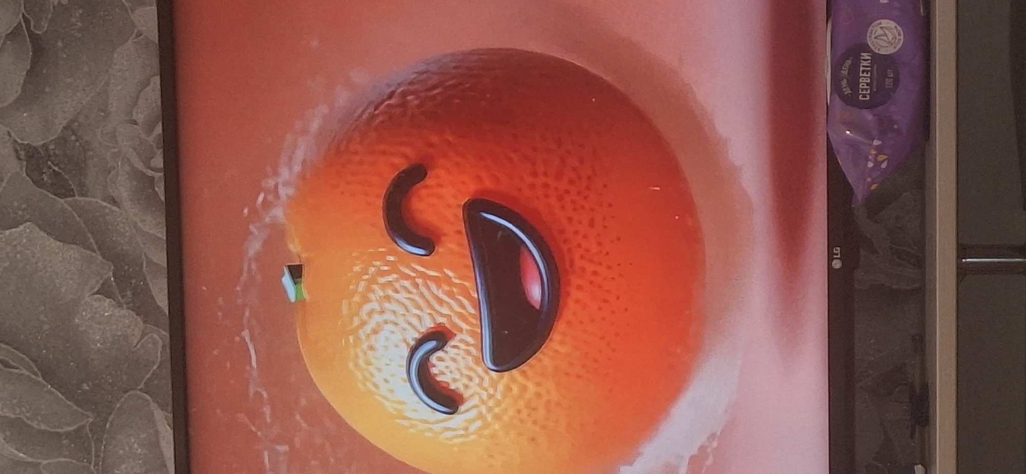 A close-up, eye-level, horizontal, full shot shows a cartoon-like orange with a happy face on a digital screen. The orange has wide, black, curved eyebrows and a large, open, black mouth with a red interior. A small green stem protrudes from the top of the orange. White, wispy elements surround the orange, suggesting a liquid or airy environment. Below the screen, a dark gray bezel of an LG television is visible, with the LG logo on the left. To the right of the logo, a portion of a purple package of wet wipes with Russian text is visible, including the word "СЕРБЕТКИ" (servetki), meaning "wipes". The background of the screen is an orangey-red gradient. The scene appears to be indoors, with the television placed on a shelf. No specific location in Bila Tserkva, Ukraine is identifiable.