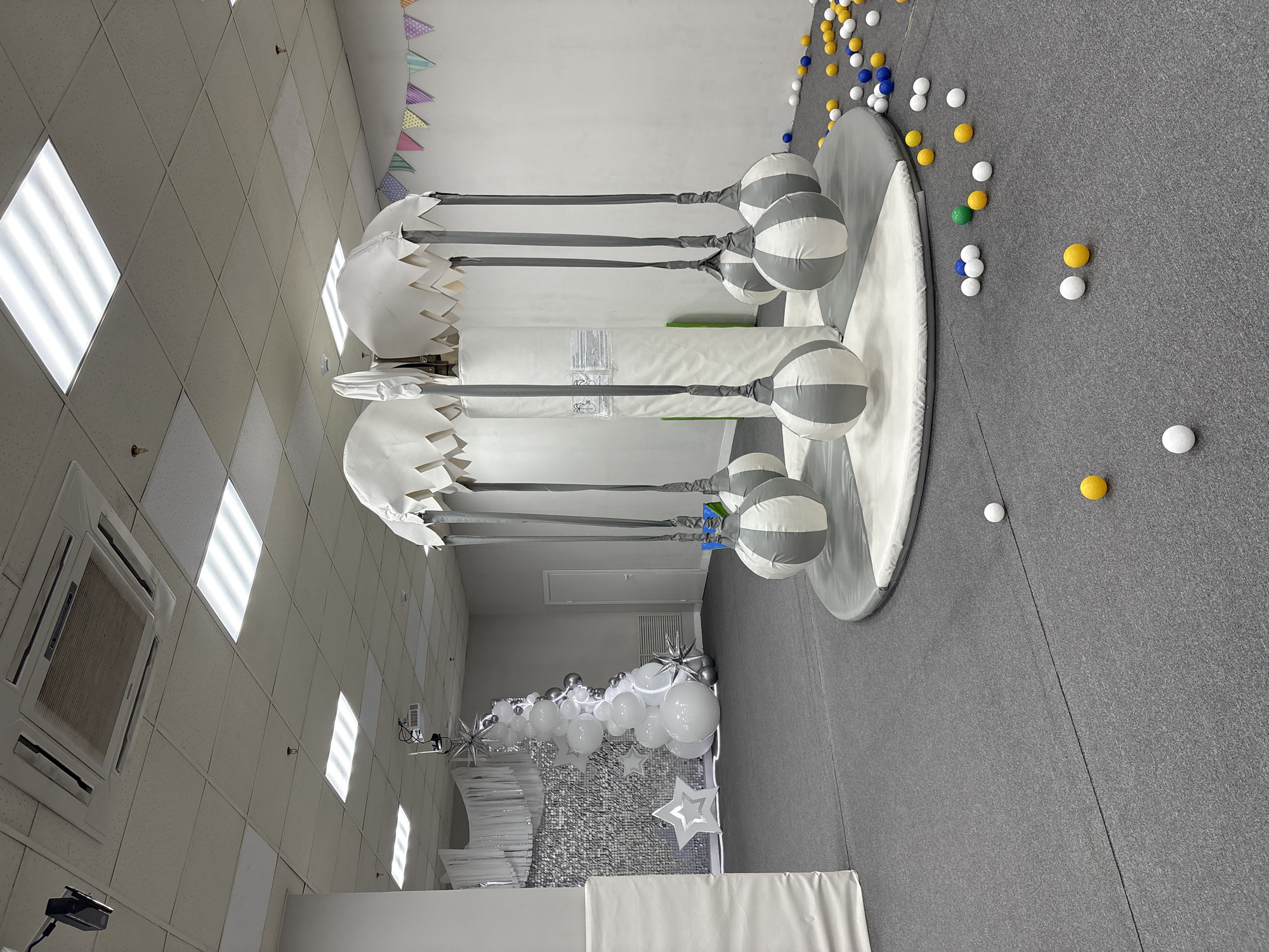 The image shows an indoor play area or children's entertainment center. The room has a grey floor with scattered balls. A circular structure with striped balls hanging from it is located in the play area. In the background, there is a decorative balloon arrangement with white and silver balloons and star-shaped decorations. The room has a white ceiling with square lighting panels.
While the image suggests a children's play area, there is no information in the image to indicate its location in Krasnoyarsk, Russia, or any city.