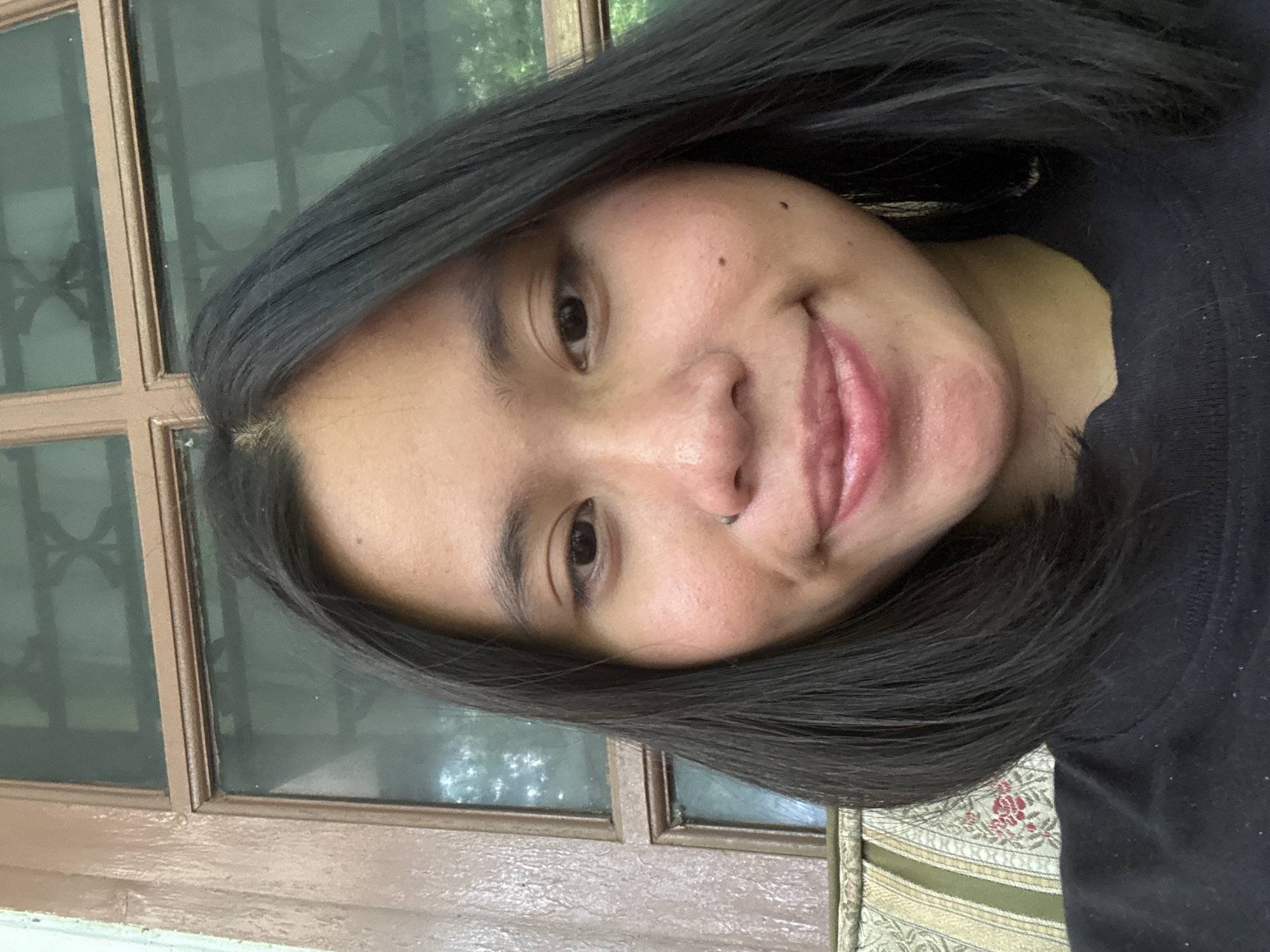 A close-up selfie shows a young Indonesian woman with shoulder-length black hair and a light brown complexion. She is looking directly at the camera with a subtle smile. She has a nose ring, two small beauty marks on her right cheek, and her lips are slightly parted. She is wearing a black t-shirt. Behind her, a wooden-framed window with glass panes is visible, with hints of green foliage and what appears to be a patterned cushion or upholstery to her left. The setting appears to be a residential area in South Jakarta, Indonesia.