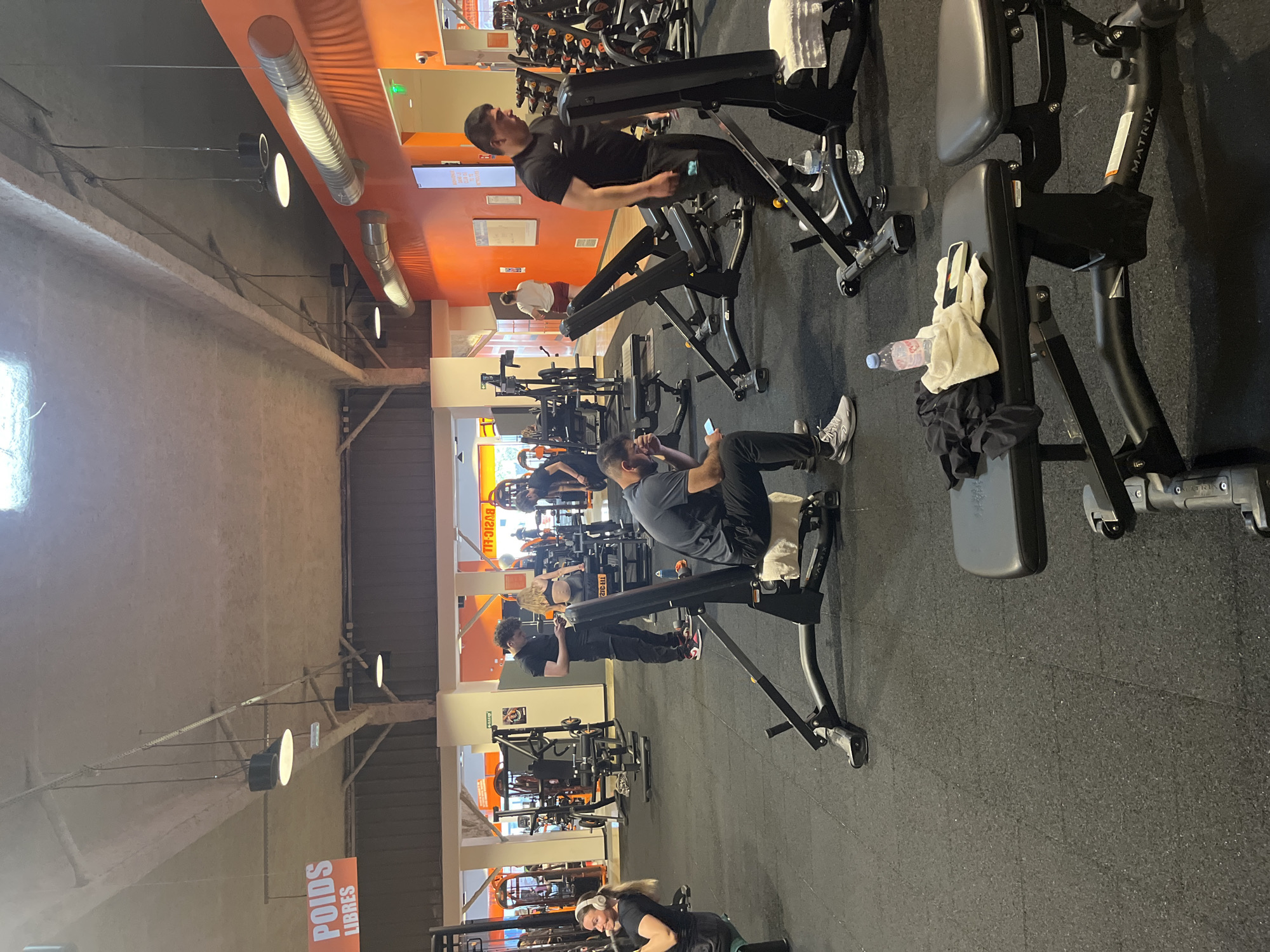The image shows the interior of a gym in Geispolsheim, France. The gym has a high ceiling with exposed beams and industrial-style lighting. Walls are painted orange and white. Several people are present, some using weight machines and others looking at their phones. The gym equipment includes various weight machines, identified by the "Matrix" brand on one machine. A water bottle and a towel are placed on a weight machine. The floor is dark-colored rubberized material. Signage in French is visible.