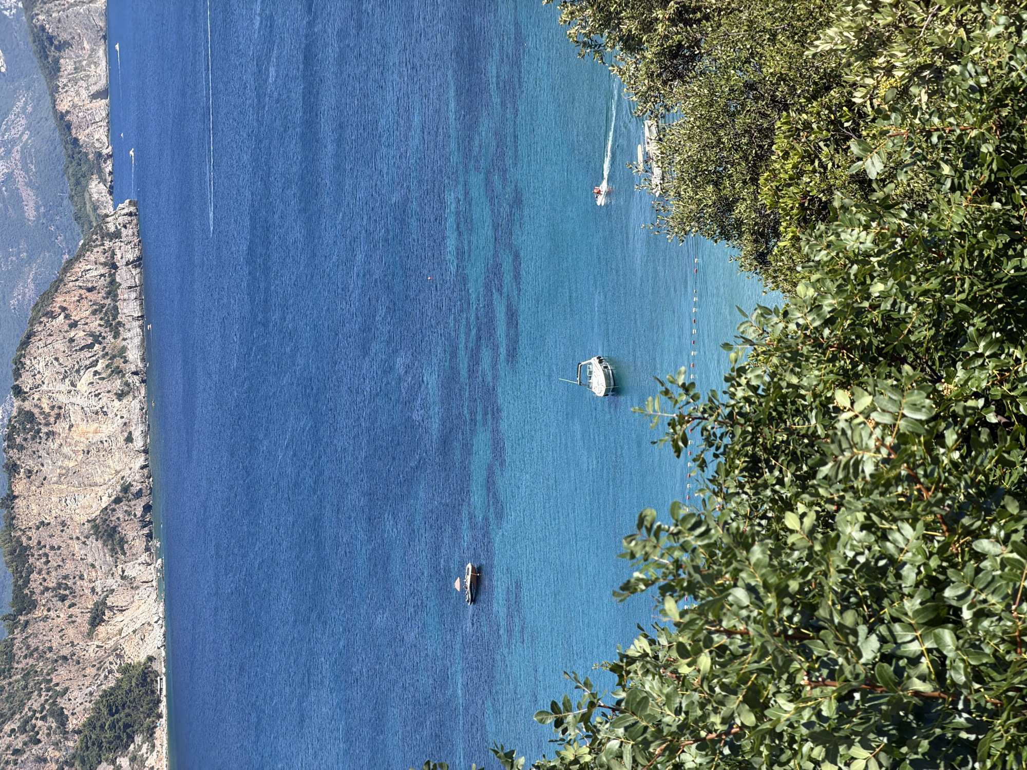 The image depicts a coastal scene in Dubraljevina, Montenegro, focusing on the azure waters of the Adriatic Sea and the rugged cliffs that frame it. In the foreground, lush green foliage partially obscures the view of the sea. Several boats are visible in the water: a small, light-colored vessel with a canopy is anchored further out, while closer to the shore, a white speedboat with two people on board is creating a wake. Another white boat is moored to the left. The clear, turquoise waters transition to a deeper blue further offshore. Rocky cliffs, dotted with sparse vegetation, rise steeply from the coastline, with a few buildings or structures visible at the base of the cliff on the left side. The overall impression is one of a serene, sun-drenched Mediterranean landscape.