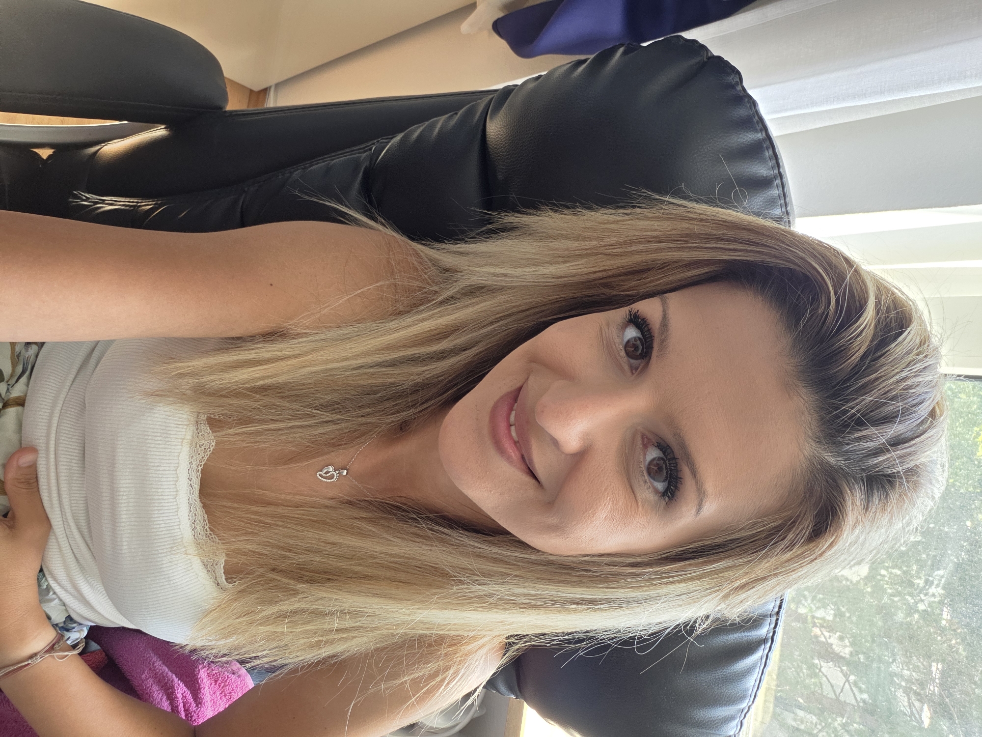 A woman with blonde hair and brown eyes is sitting in a black office chair, looking at the camera. She is wearing a white tank top with lace trim and a silver necklace with a heart and footprints pendant. The chair is positioned in front of a window with white curtains. Outside the window, trees are visible. The room appears to be an office or a home office. The woman's left arm is resting on her stomach, and her right hand is also visible near her stomach. A pink cloth or blanket is draped over the back of the chair to her right. The lighting suggests it is daytime, possibly in the late morning or early afternoon. The location is not specified as Munich, Germany, in the provided image.
