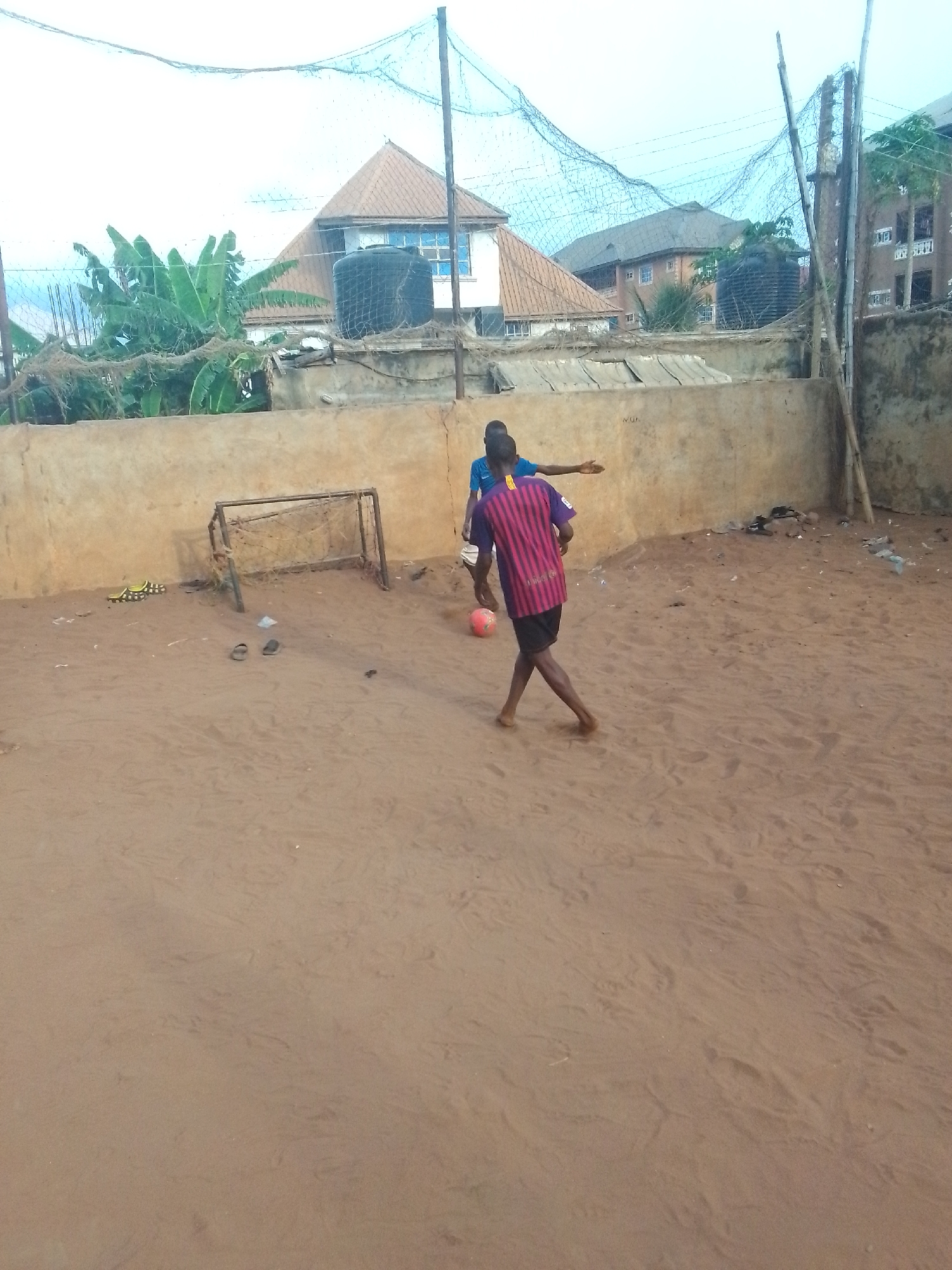 In an outdoor dirt field, two people are playing soccer near a small metal goal. One person wears a striped shirt and black shorts, and the other wears a blue shirt. A red soccer ball is between them. Behind them is a low, light brown wall, and above that are buildings, water tanks, and netting supported by poles. There is an object lying in the dirt on the left side of the scene.