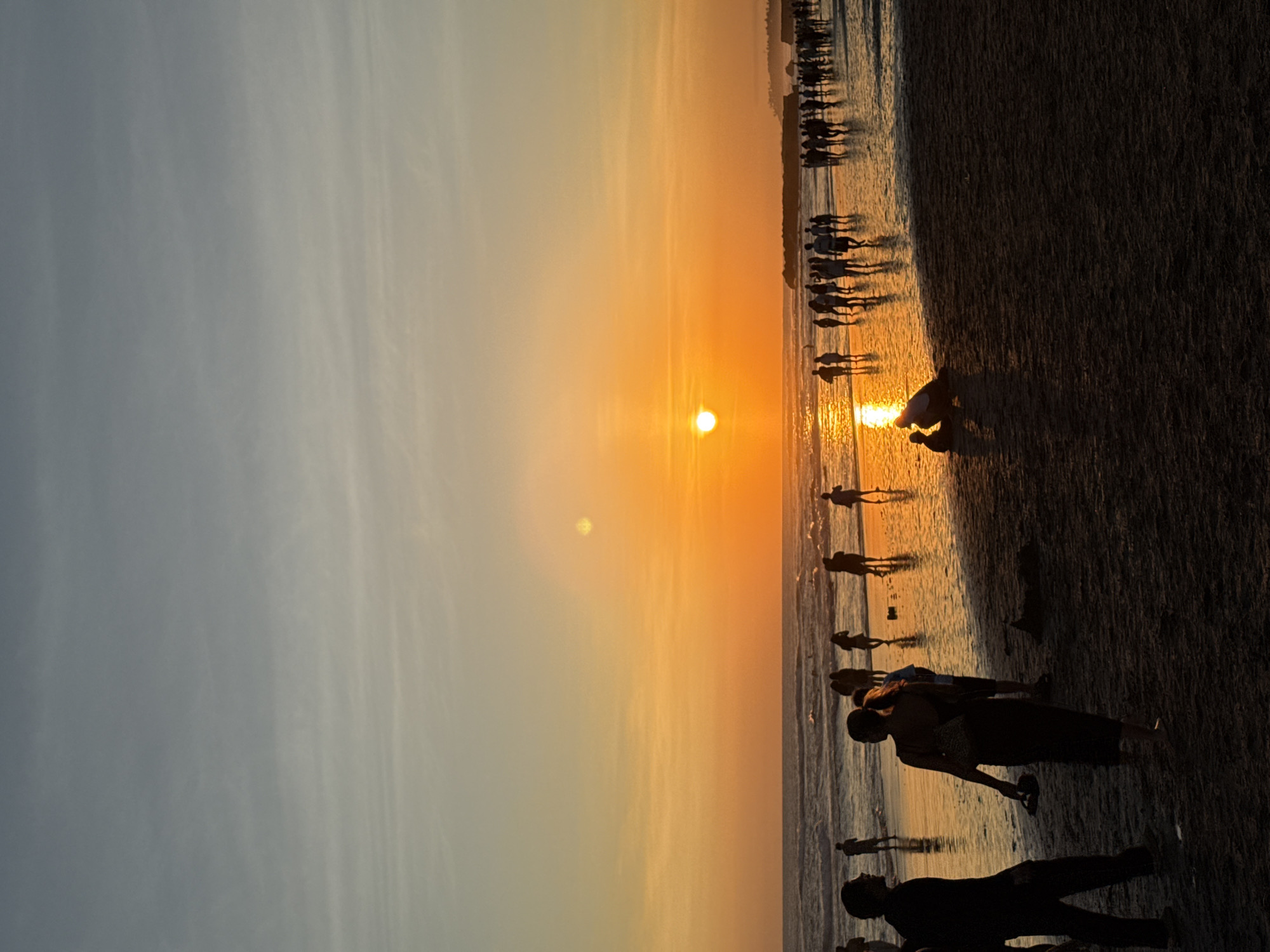 The image depicts a sunset on a beach in North Kuta, Indonesia. The sun is setting on the horizon, casting an orange glow on the sky and reflecting on the water. Silhouettes of people are visible along the shoreline, some walking, standing, and interacting. The beach is a dark color, contrasting with the bright sky.