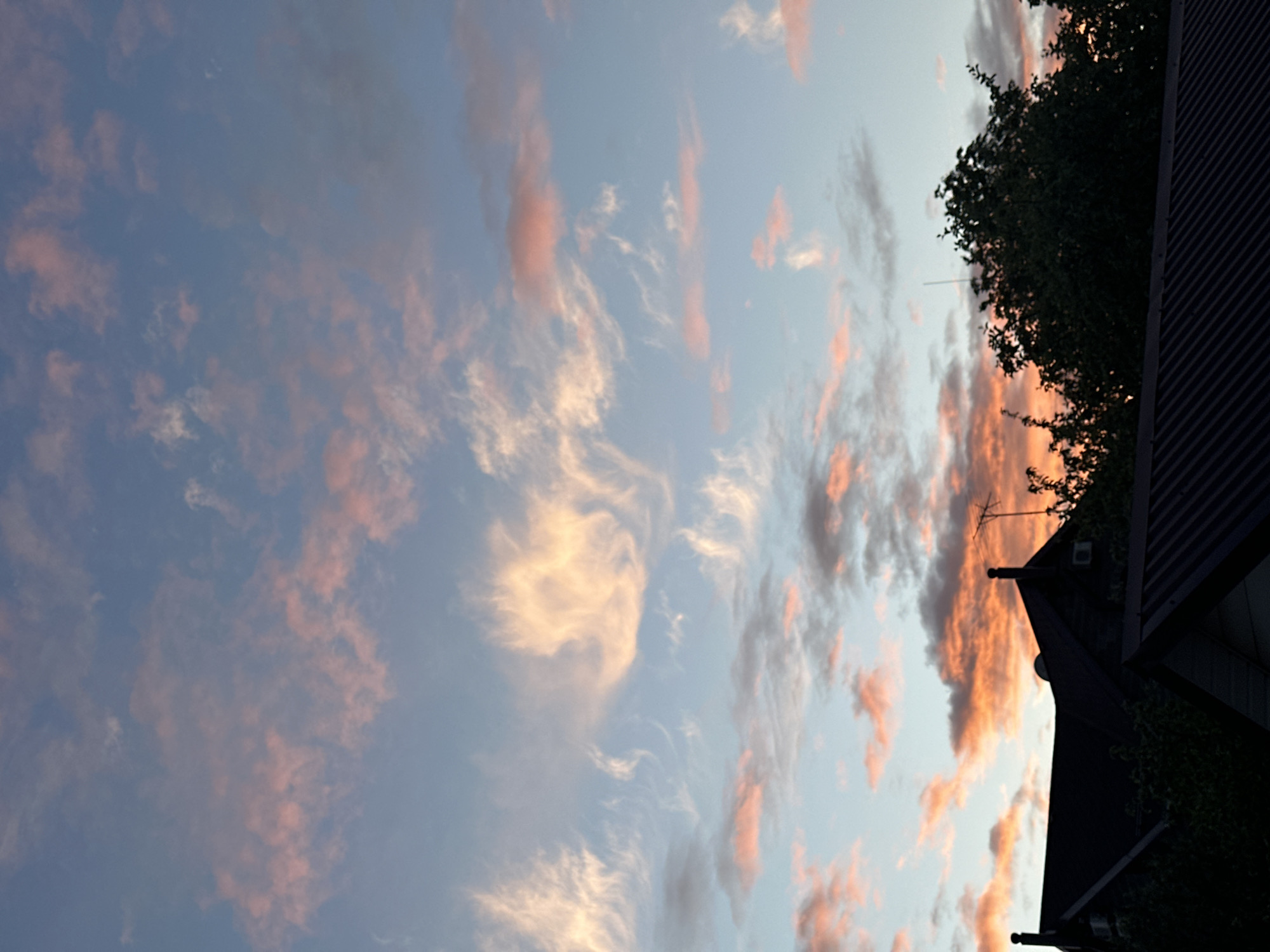 The image captures a view of the sky at dusk, with clouds colored in hues of pink and orange by the setting sun. A portion of a building is visible on the right side of the frame. The location is not discernible from the image. No people or specific actions are observable.