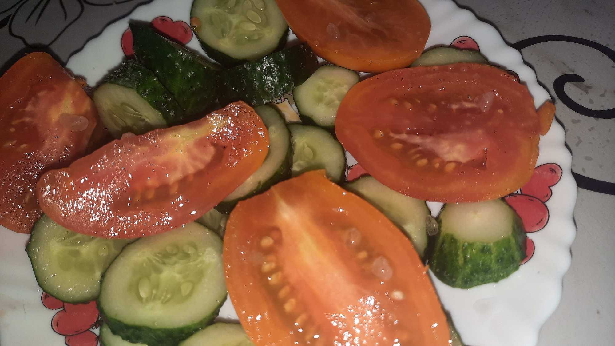 A close-up, overhead view captures a portion of a white, scalloped plate filled with sliced tomatoes and cucumbers. The vegetables are arranged haphazardly, with some slices overlapping. The tomatoes are bright red and appear juicy, with visible seeds and pulp. The cucumbers are a vibrant green on the outside with lighter green flesh and visible seeds on the inside. The plate's rim features a pattern of red heart-like shapes. The background is out of focus and appears to be a gray surface. The scene is likely from a home kitchen in Shklov, Belarus, given the context provided.