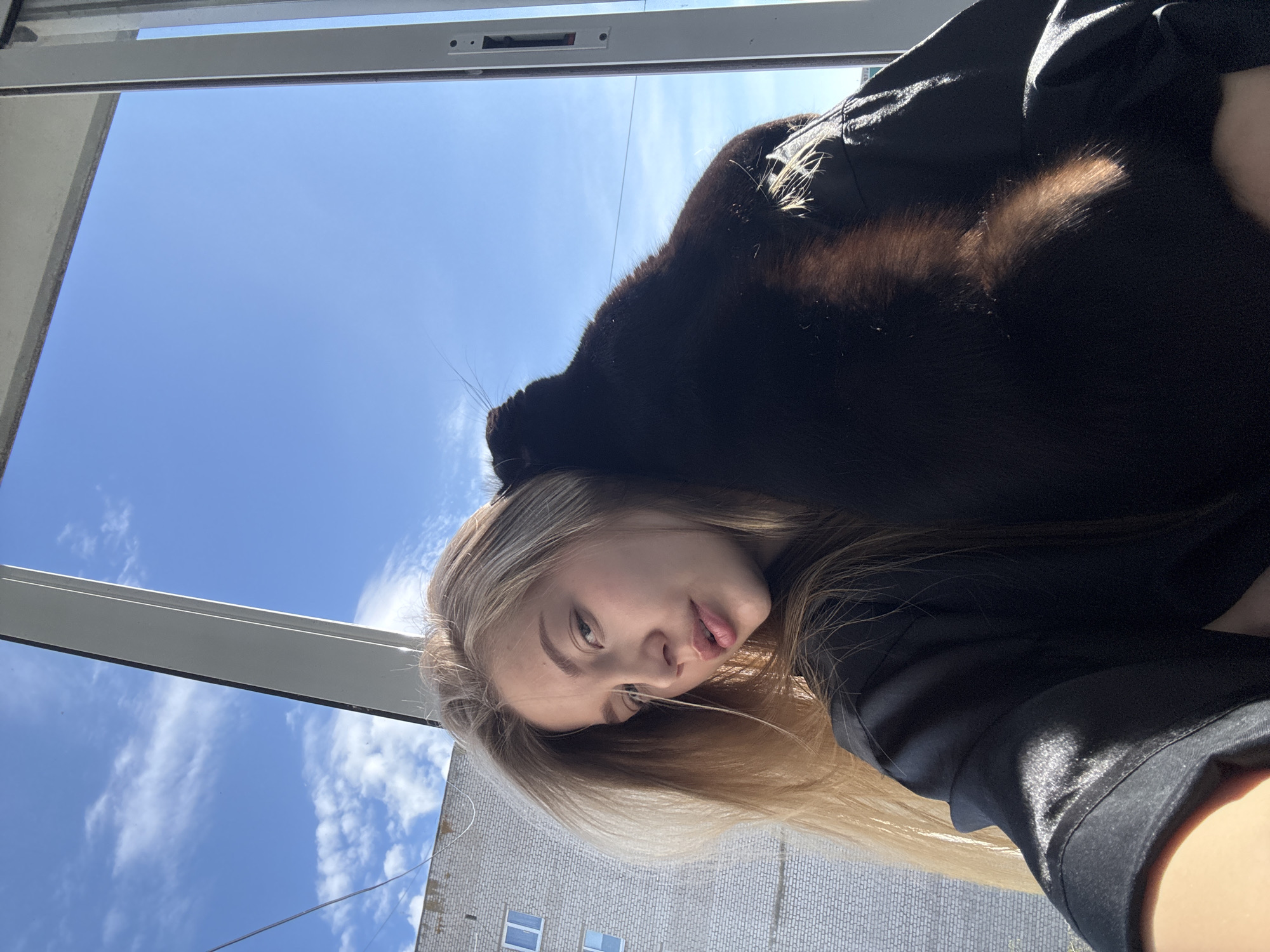 This media file captures a close-up portrait of a young woman with a black cat, taken indoors looking out through a window. The woman has long, straight blonde hair and is looking directly at the camera with a subtle pout. A black cat is positioned on her head and shoulder, with its whiskers clearly visible and some of its fur illuminated by direct sunlight, showing hints of brown or reddish tones.
The setting outside the window reveals a bright, clear blue sky with sparse white clouds, indicative of a sunny day, likely during midday or afternoon. A white window frame is prominent in the upper and left portions of the image, framing the outdoor view. Below the sky, part of a building with a light-colored, brick-like texture and several windows can be seen, suggesting an urban or suburban residential area. The overall scene conveys a relaxed moment between the woman and her pet. No visible text is present in the image. The location context for this scene is Chaikovsky, Russia.