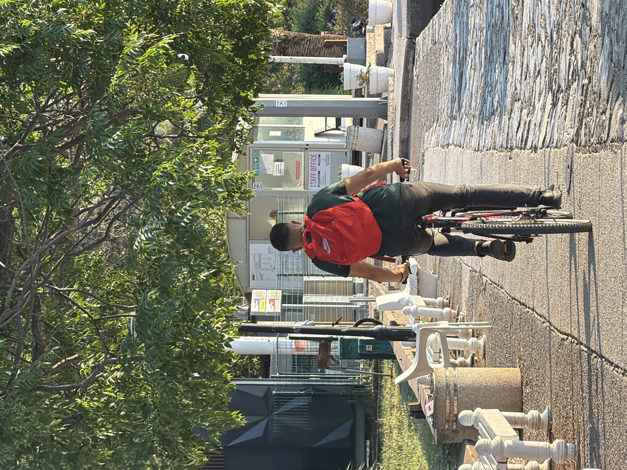 A person wearing a dark green t-shirt, dark pants, and black shoes is riding a red and black bicycle. They have a red backpack on their back. The person is cycling on a paved path that appears to be made of cobblestones. To the left of the path, there is a grassy area with white benches and a cylindrical trash can. In the background, there is a small building labeled "STAFF OFFICE" with a fence in front of it. Several white potted plants are visible near the building. Trees with green foliage hang over the scene, casting shadows on the path. The lighting suggests it is daytime, possibly late afternoon due to the long shadows. This scene appears to be in a public area, possibly a promenade or park, adjacent to a body of water that is not directly visible but implied by the waterfront setting. The location is the promenade in front of the "Staff Office" building of the Stari Grad (Old Town) area in Budva, Montenegro.