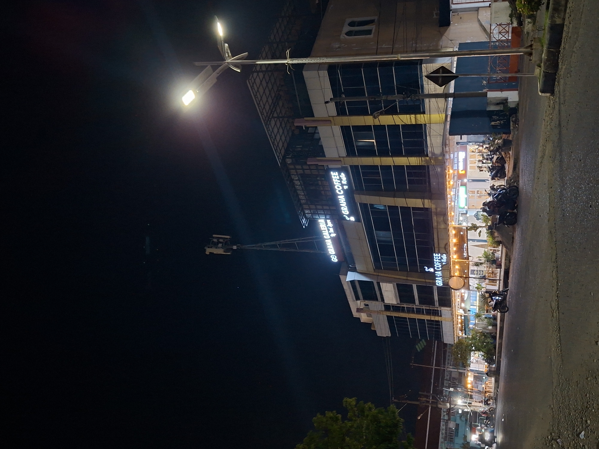 At night in Lhokseumawe, Indonesia, a street is lit by streetlights and neon signs. The multi-story Graha Coffee shop displays brightly lit windows and signs. Parked motorcycles line the sidewalk. The street surface appears wet. A communication tower rises in the background. A few people are visible in the distance.