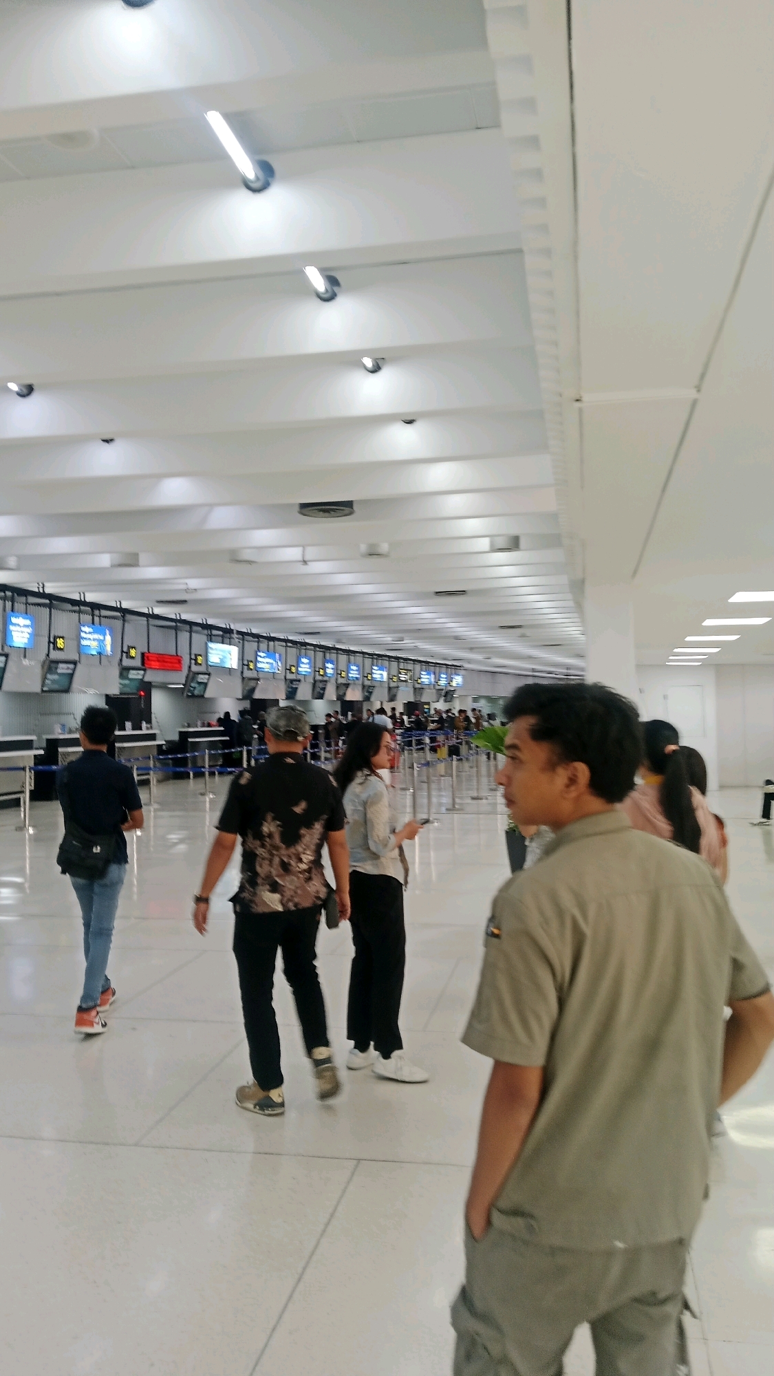 The image depicts the interior of an airport terminal, likely located in Tangerang, Indonesia, given the context. The scene is brightly lit by fluorescent lights mounted on the white, slatted ceiling. In the foreground, a person in a tan uniform is facing away from the camera, with their hands in their pockets, and appears to be standing. Behind them, several other individuals are walking through the terminal. To the left, two men are walking towards the check-in counters, one wearing a dark shirt and jeans with a black bag slung across his body, and the other in a patterned black shirt and dark pants. Between them, a woman in a light-colored shirt and dark pants is walking while holding a phone. In the background, a long row of check-in counters with digital displays is visible, with additional people queuing. The floor is a polished, light-colored surface.