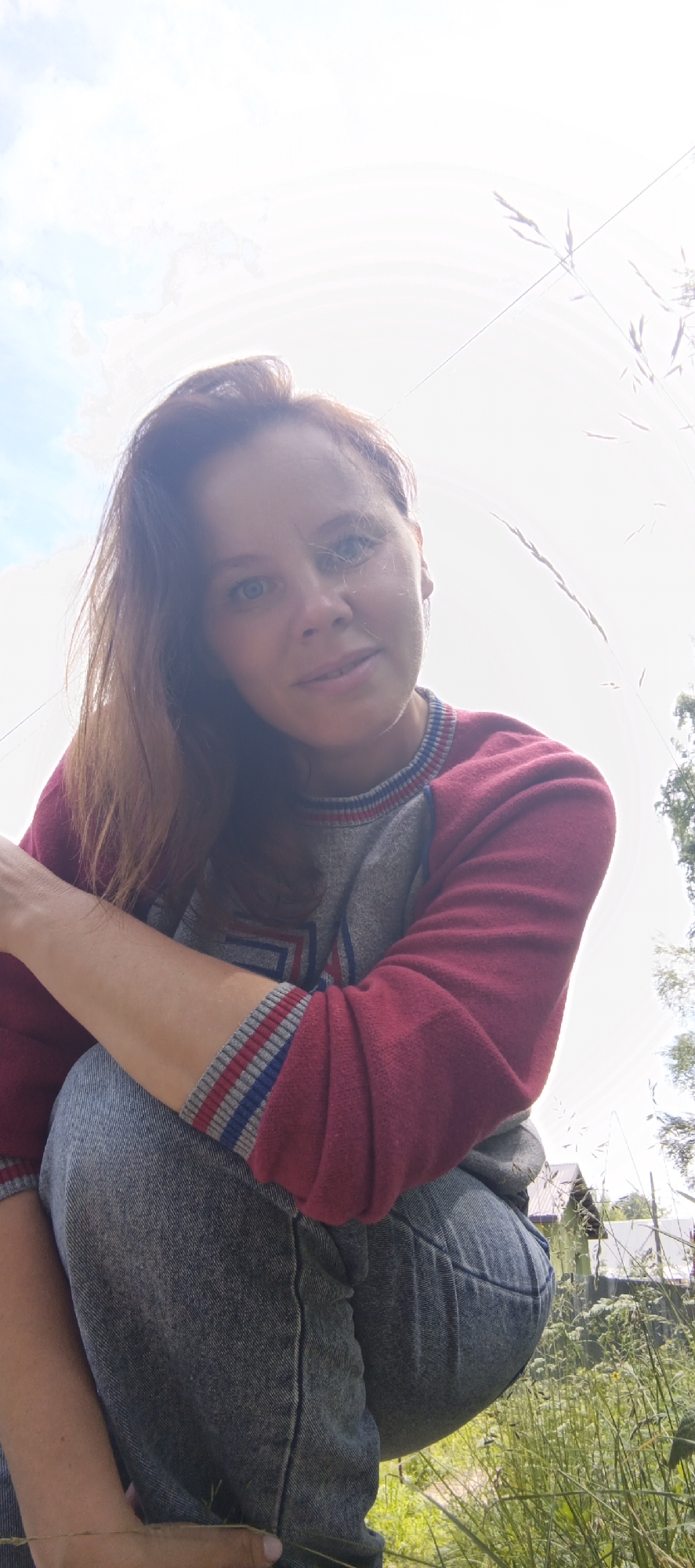A medium shot captures a woman with auburn hair and blue eyes. She is wearing a maroon and gray sweatshirt with striped cuffs and blue jeans. The woman is crouched down, looking towards the camera, with her left arm resting on her bent knee. The background is a bright sky and some blurry green foliage, suggesting an outdoor setting, possibly in a rural or suburban area. The image is taken from a low angle.