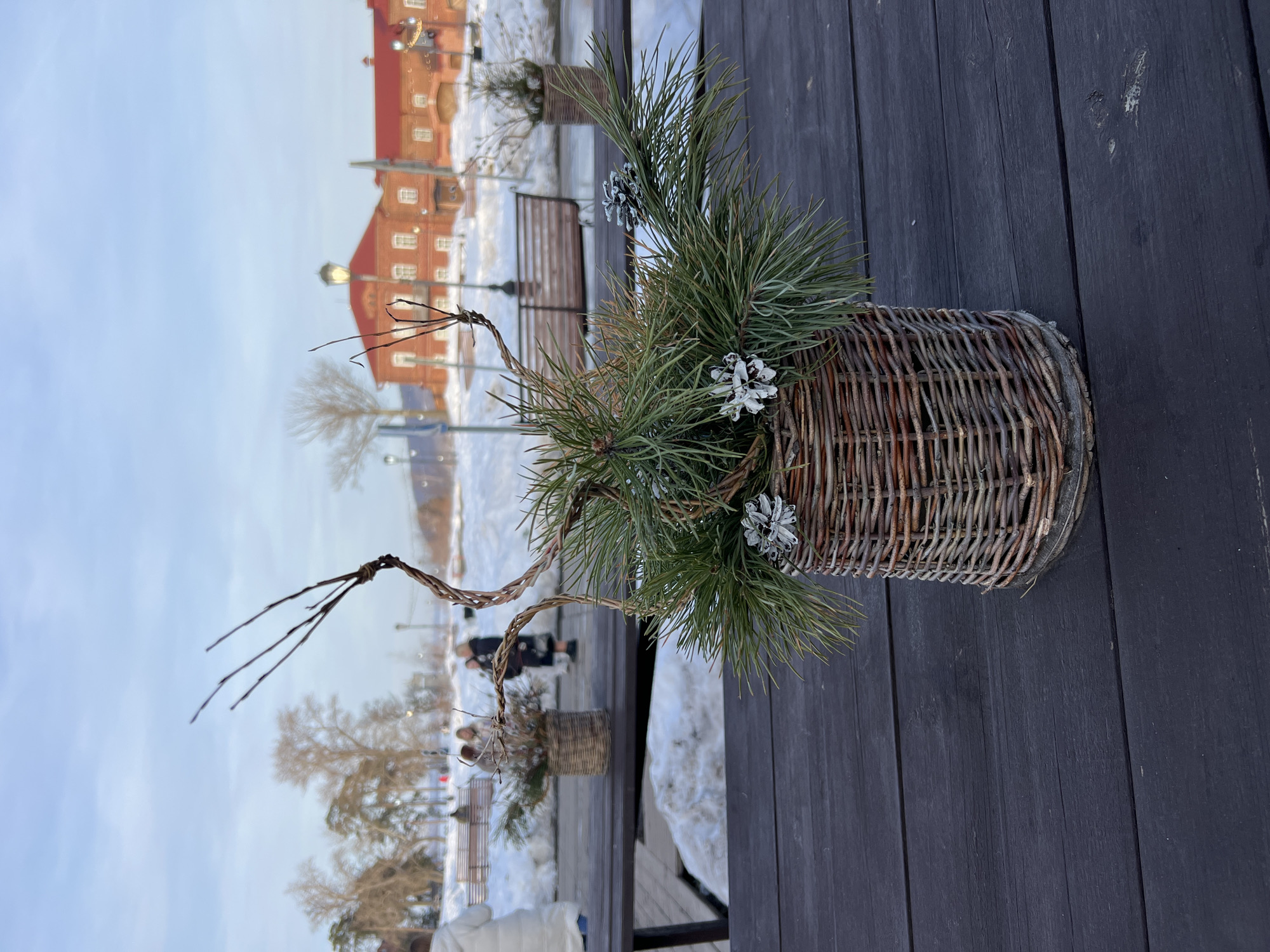 A rustic wicker basket, approximately cylindrical, sits slightly right of center on a dark, weathered wooden plank surface. The basket contains an arrangement of green pine branches, two small frosted white pine cones, one white snowflake-shaped ornament, and several bare, dark brown, intertwined branches extending upwards. In the blurred mid-ground, a similar smaller wicker basket arrangement is visible on a lower wooden platform to the left. The background reveals a snow-covered landscape in the city of Sviyazhsk, Russia. Several indistinct human figures in winter attire are present, walking or standing amidst the snow. Wooden park benches are partially visible. Further back, red-roofed, light-colored brick buildings with multiple windows and a black streetlamp are discernible. Bare deciduous trees and evergreen trees dot the landscape. The sky is a clear, light blue, indicating daylight conditions. Another wicker basket decoration is visible on a bench in the far right background.