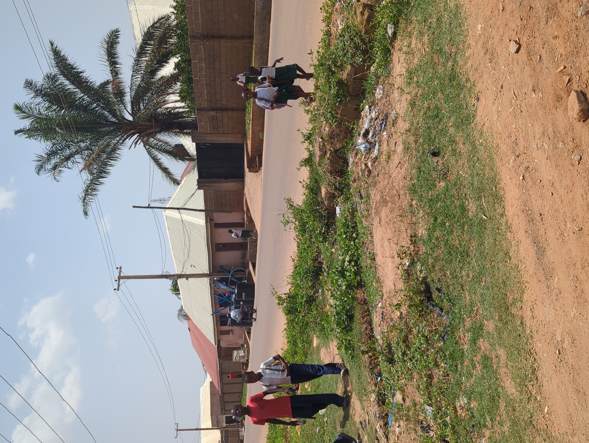 The image depicts a street scene in Narayi, Nigeria. The foreground features a dirt shoulder transitioning to a paved road. Two men stand on the shoulder, one wearing a red shirt and the other a white shirt with blue jeans. Further down the road, three schoolgirls in green skirts and white shirts are walking. In the background, buildings line the street, including a structure with a light-colored roof. A palm tree stands prominently on the left side. Power lines run above. Various objects are present, including a bicycle parked near one of the buildings. Some trash is strewn along the roadside. The weather appears sunny.