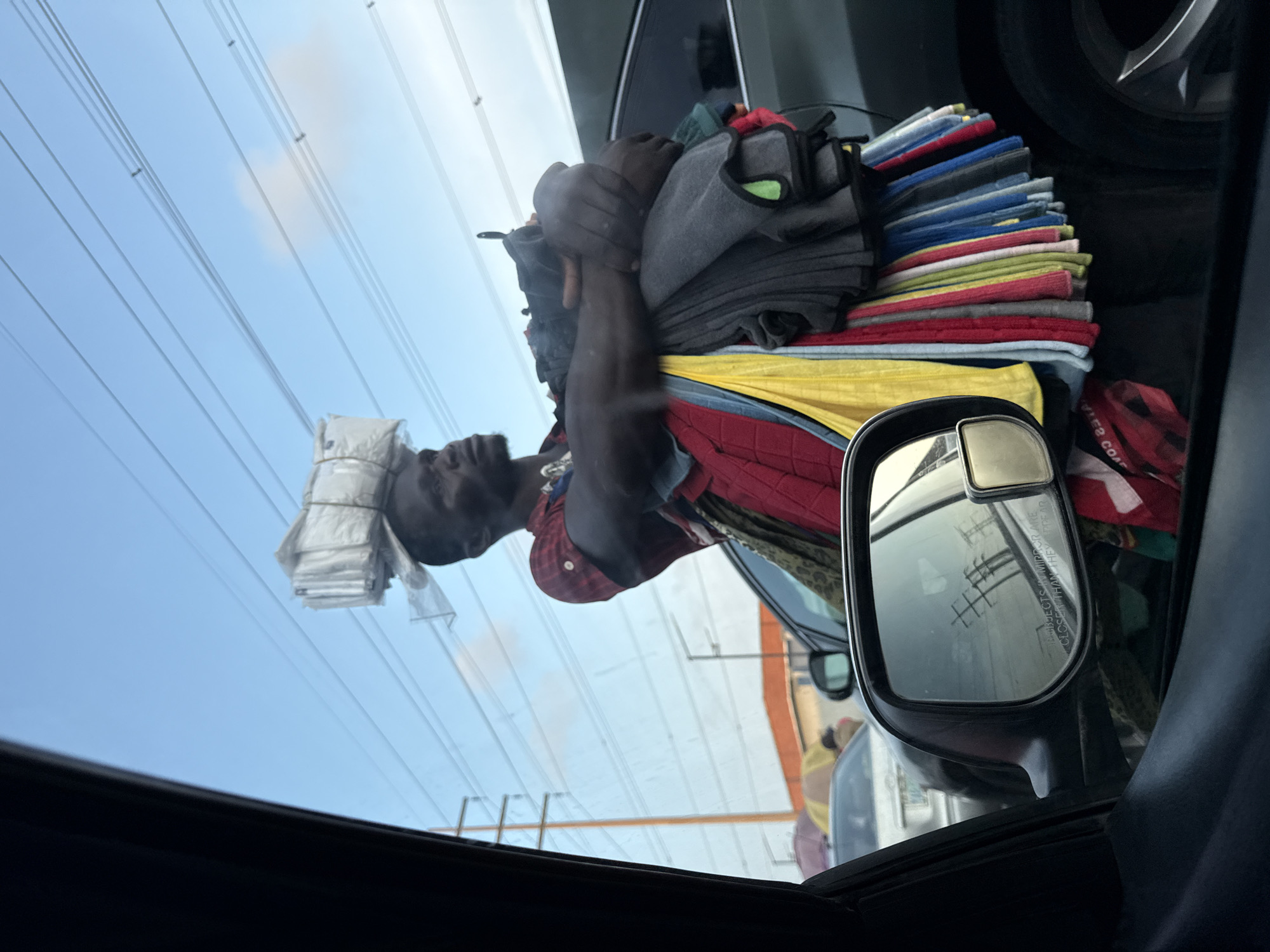 A man, likely a vendor, stands on a roadside in Lekki, Nigeria. He is carrying a large stack of white plastic-wrapped packages on his head, secured with twine. He is also holding a large bundle of colorful cloths, predominantly red, yellow, gray, and blue, in his arms. He appears to be standing between vehicles, possibly in traffic. The background shows power lines and utility poles, a street light, and buildings in the distance. The scene is captured from inside a vehicle, as evidenced by the car's side mirror and window frames in the foreground. The sky is clear blue with a few scattered clouds.