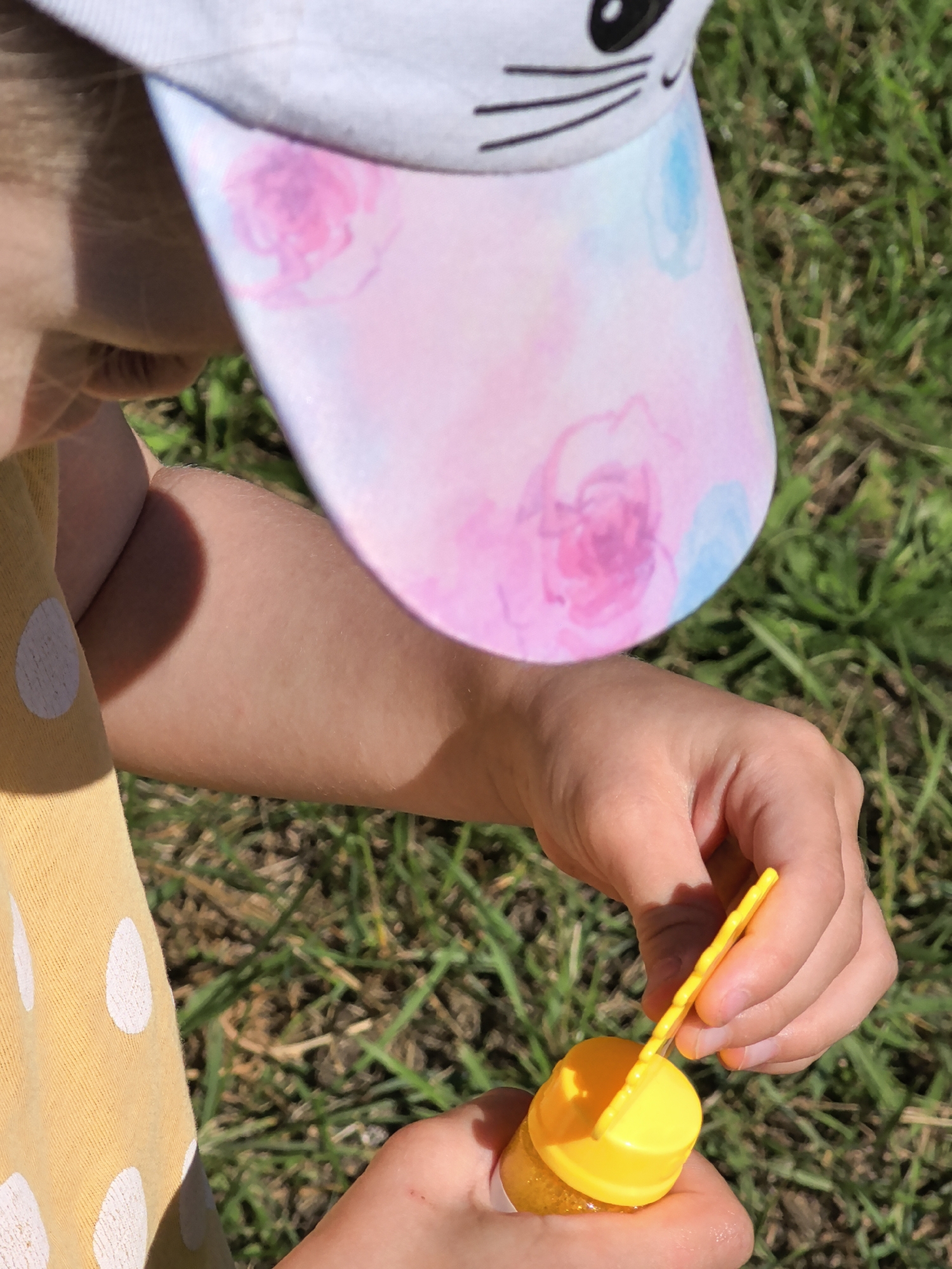 A close-up shot captures a child's hands engaged in blowing bubbles outdoors. The child wears a yellow shirt with white polka dots and a white baseball cap decorated with a cartoon cat face and watercolor-style pink and blue floral patterns. One hand holds a small yellow bottle of bubble solution, while the other inserts a yellow bubble wand into the bottle. The background consists of green grass, suggesting a daytime scene on a sunny day. The focus is on the activity of bubble blowing and the details of the child's attire. No visible text is present in the image.