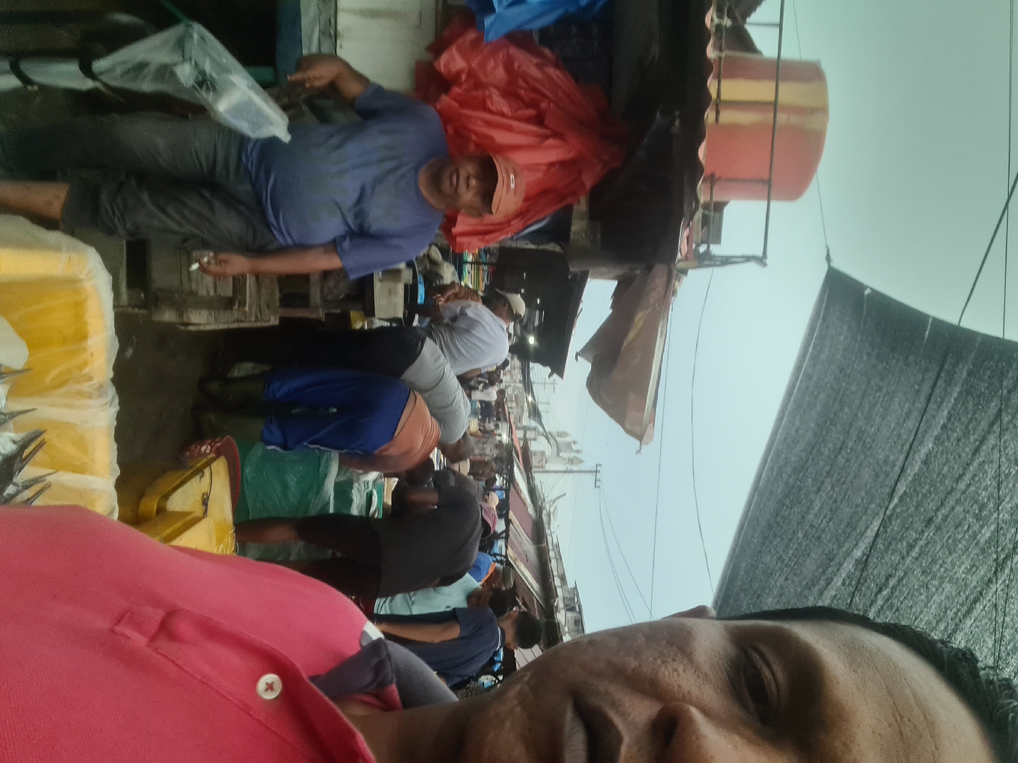 The image shows a street market scene, likely the traditional fish market in Lhokseumawe, Indonesia. In the foreground, a man is taking a selfie, with his face close to the camera on the right side of the frame. He is wearing a pink polo shirt. In the background, various market stalls are visible, with people engaged in activities typical of a market. One stall has a large red plastic covering. A man in a blue t-shirt and a cap is standing near some plastic bags, while other individuals are working with goods at different stalls. The environment appears to be outdoors, under a partially covered area, suggesting it is a bustling market setting. The sky is overcast.