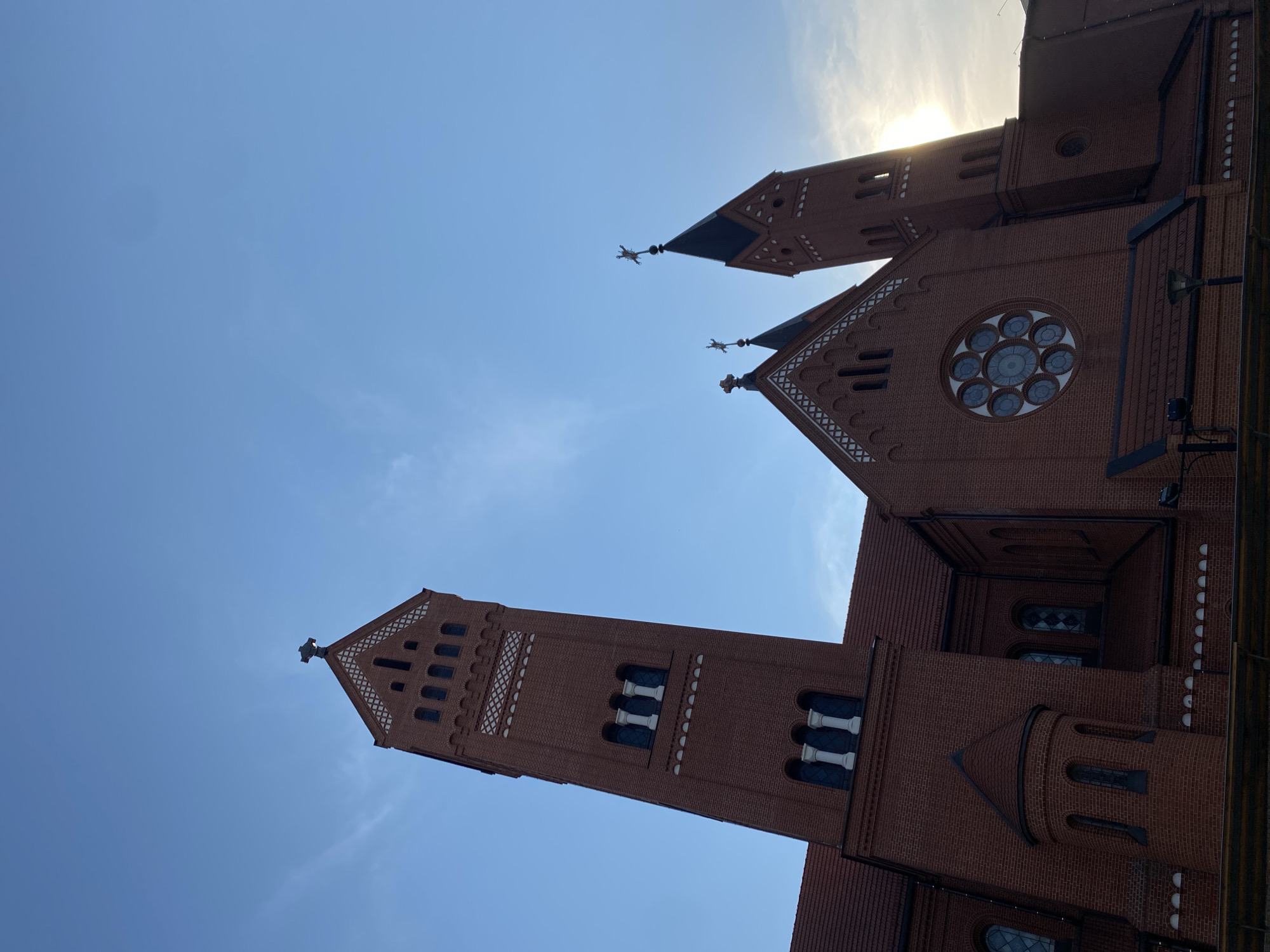 This low-angle, wide shot captures the red brick facade of the Church of Saints Simon and Helena in Minsk, Belarus. The sky is a clear, bright blue with some wispy clouds and the sun is shining through the upper right portion of the frame, indicating it's likely daytime. The church features two prominent towers, each topped with a pointed roof and decorative elements. One tower is in the foreground on the left, and another is further back on the right. The building has gothic architectural elements, including pointed arches, decorative window tracery, and a large rose window on the main building. The overall impression is one of grandeur and architectural beauty against a clear sky. No people are visible in the image.