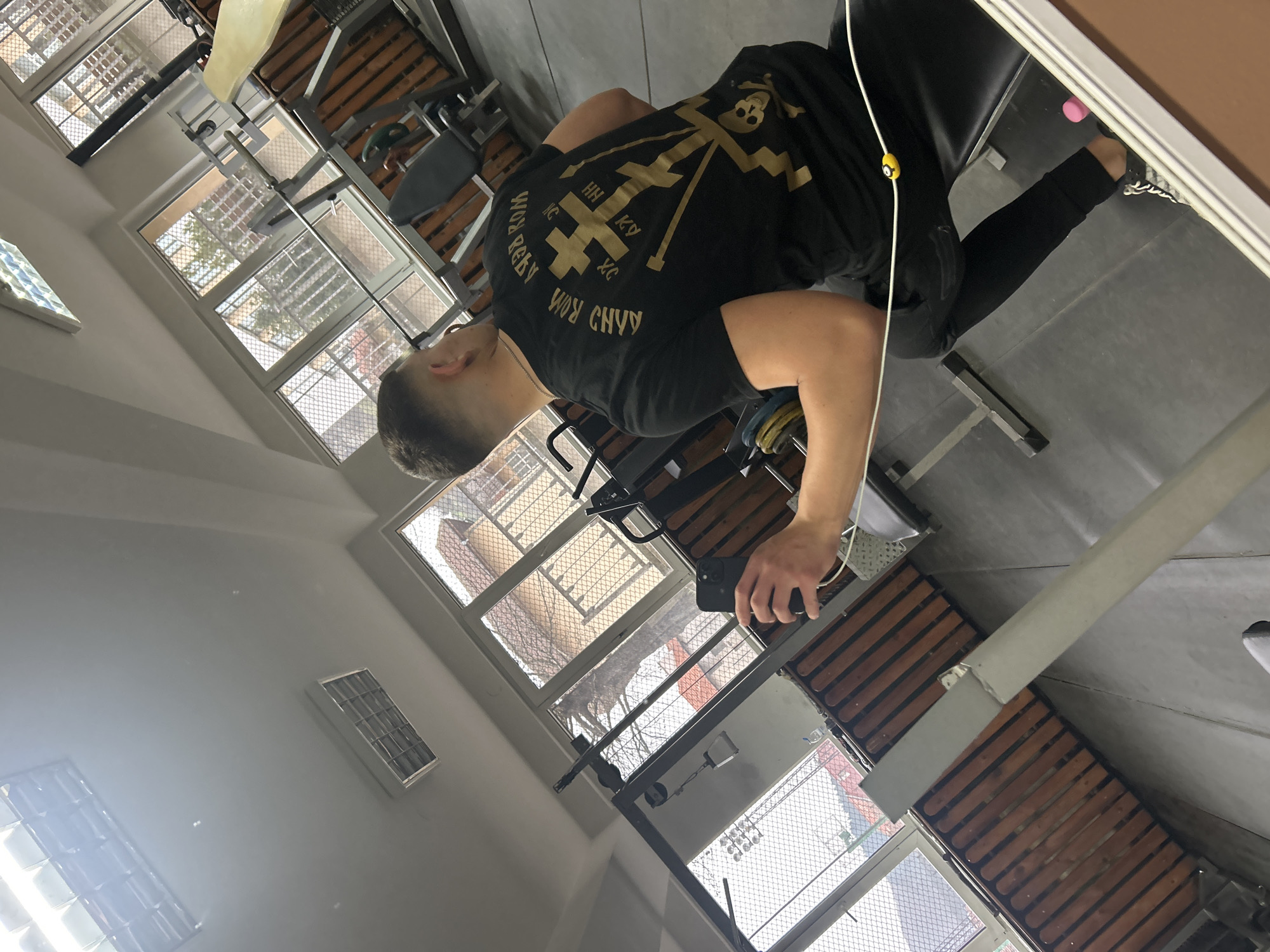 A man wearing a black t-shirt with gold-colored designs is sitting on a weight bench indoors. He appears to be holding a phone, possibly taking a picture of himself in a mirror. The room contains exercise equipment and large windows with wire mesh, offering a view of buildings outside. The precise location within Zlatograd, Bulgaria cannot be determined from the given image.