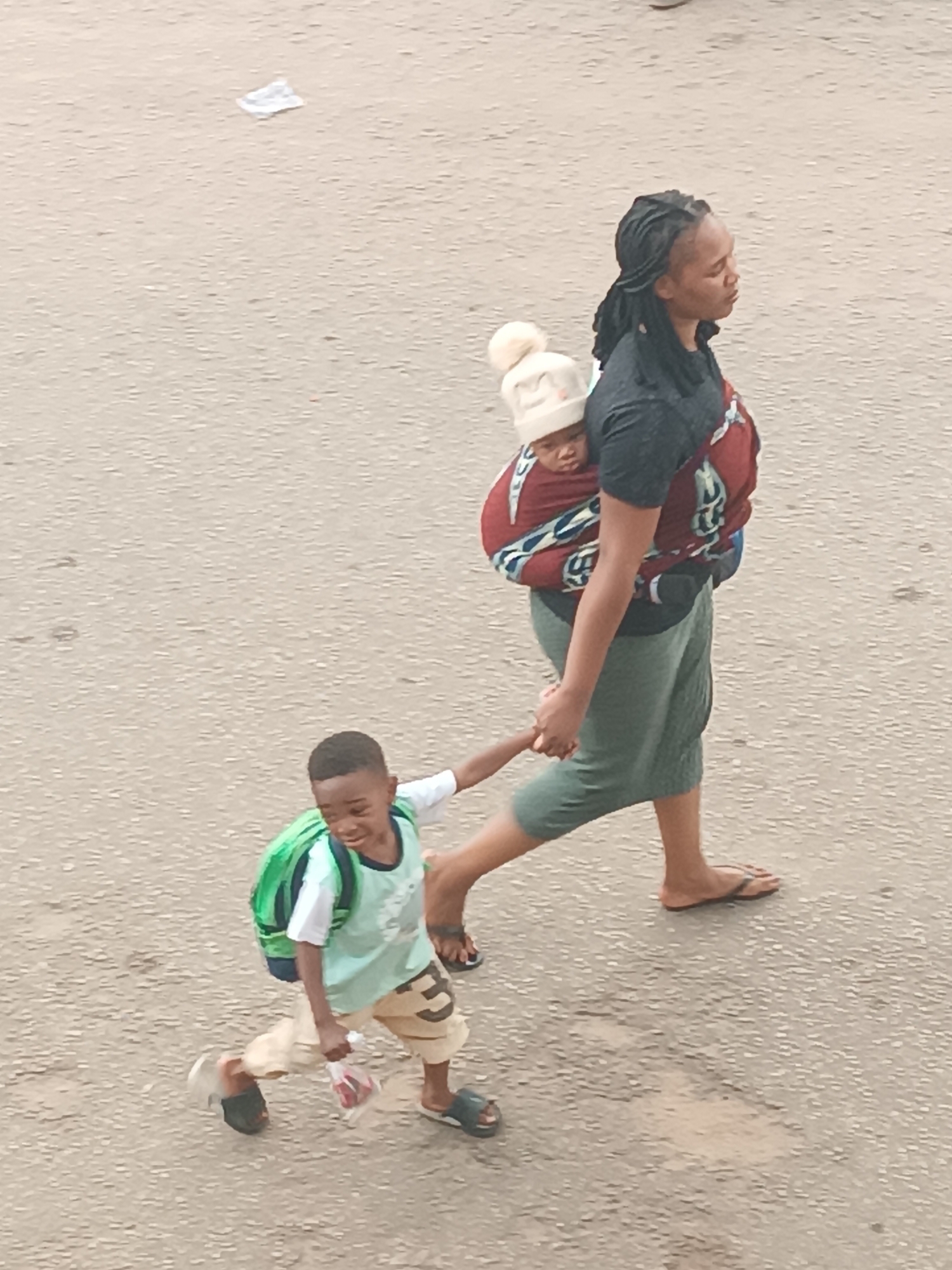An overhead shot captures a woman walking with two children on an outdoor, textured, light-colored asphalt or concrete surface. The scene is brightly lit by natural daylight.
The woman, who appears to be an adult with dark skin and dark braided hair, is positioned on the right side of the frame, walking towards the right. She wears a dark short-sleeved t-shirt, a long green skirt, and dark flip-flops. On her back, she carries a baby securely wrapped in a red fabric sling with blue and white patterns. The baby wears a light-colored knitted hat with a prominent white pom-pom on top, and its face is visible, looking towards the left.
To the woman's left, a young boy walks energetically, holding her hand. He has dark skin and short dark hair. The boy wears a light green short-sleeved t-shirt, light brown shorts with a large dark number "3" visible on the right leg, and black flip-flops. He carries a bright green backpack and holds a small clear plastic bag containing red items in his left hand. His facial expression suggests liveliness, with a slight smile or open mouth, as he looks forward and slightly upwards.
The ground surface appears to be a well-worn road or path, with some minor debris visible in the upper left corner. The image captures a moment of everyday activity, showing a mother caring for her children while walking.