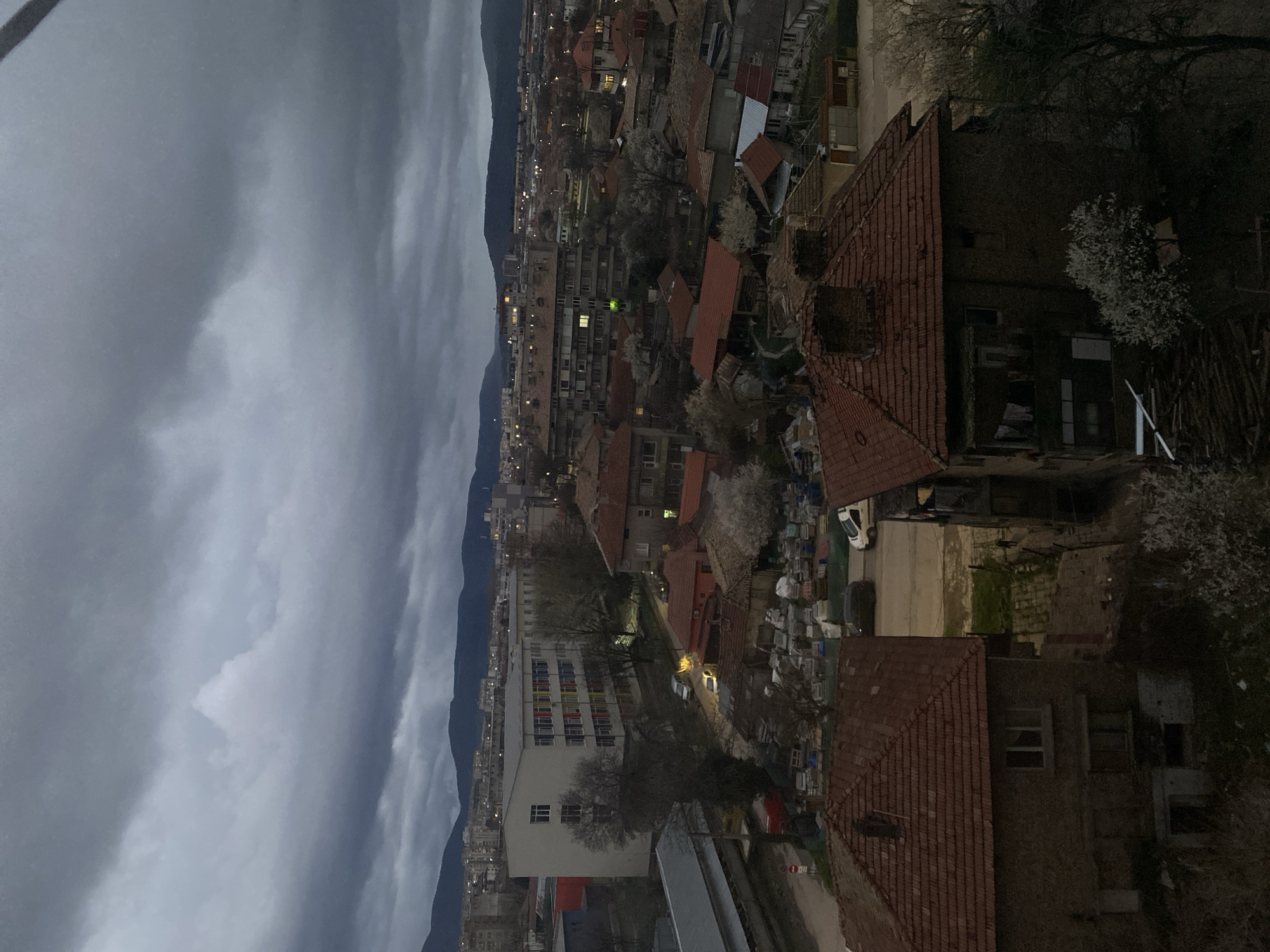 Here is a detailed description of the image:
The image presents an elevated, slightly tilted, panoramic view of an urban landscape, set against a dramatic, cloudy sky. The city is nestled against a backdrop of dark, mountainous terrain.
The city itself is composed of a mix of residential and commercial buildings. Many of the buildings have red-tiled roofs, common in this region. There is a visible contrast between older, traditional structures and more modern apartment complexes, reflecting different eras of urban development. Some buildings feature visible windows, with a few emitting light, indicating the time is likely dusk or night.
In the foreground, closer to the viewpoint, are the tops of buildings with red-tiled roofs and brick or stone facades. Trees with budding white flowers are also visible, suggesting the season is spring.
The sky is dominated by dark, heavy clouds, casting a somewhat somber tone over the scene. The mountains in the background provide a natural boundary to the cityscape.
Based on the prompt, the location is Targovishte, Bulgaria. The image captures the city's urban sprawl, nestled against the natural backdrop of the mountains, under a dramatic sky.