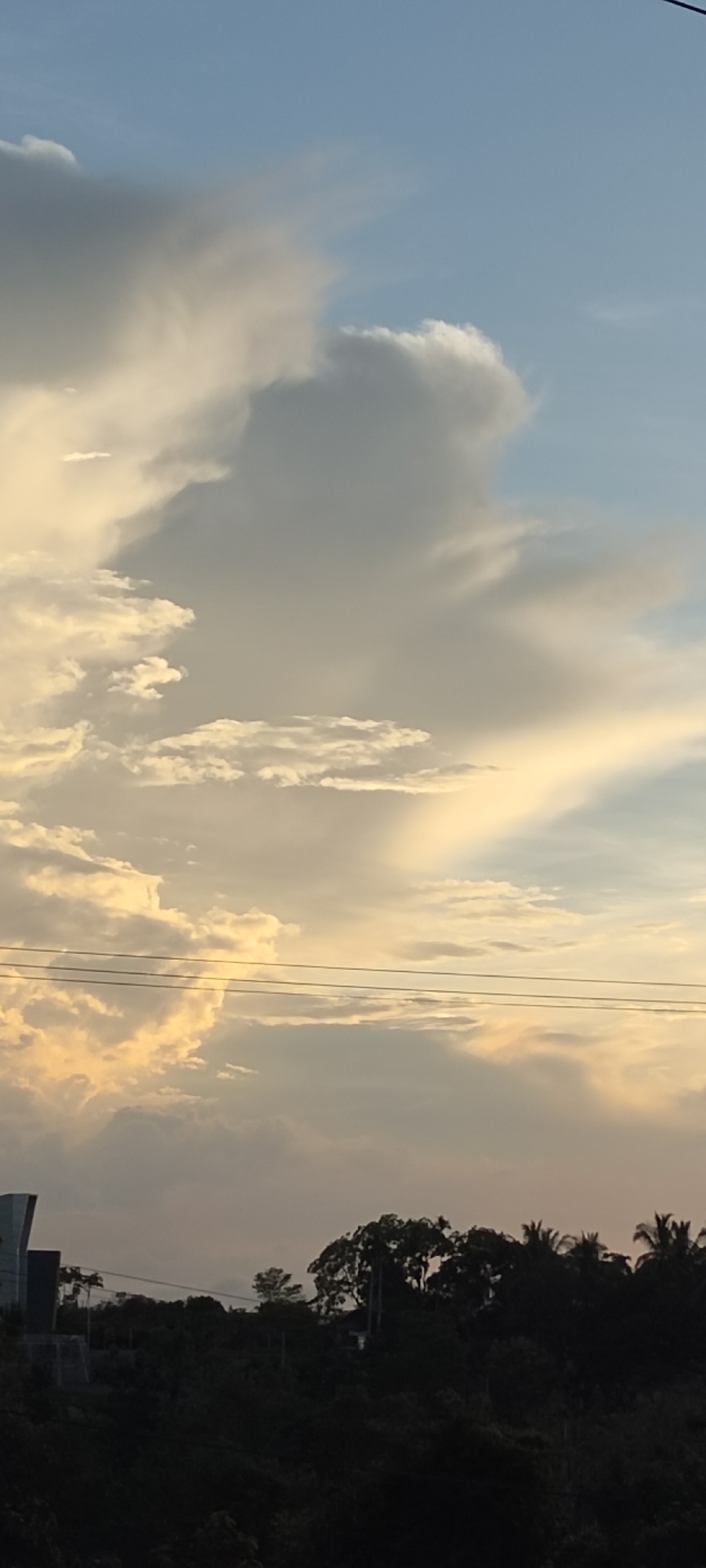 The image shows a view of the sky in Lhokseumawe, Indonesia, at what appears to be dusk or dawn. The sky is predominantly light blue, transitioning to a yellowish or golden hue closer to the horizon, indicative of sunlight filtering through the atmosphere. Various cloud formations are visible, ranging from dense, gray masses to thinner, wispy streaks. In the lower portion of the image, there's a dark silhouette of trees and vegetation, presumably forming part of the urban landscape of Lhokseumawe. Some utility lines are visible against the sky. Part of a building's structure is on the left side of the image.