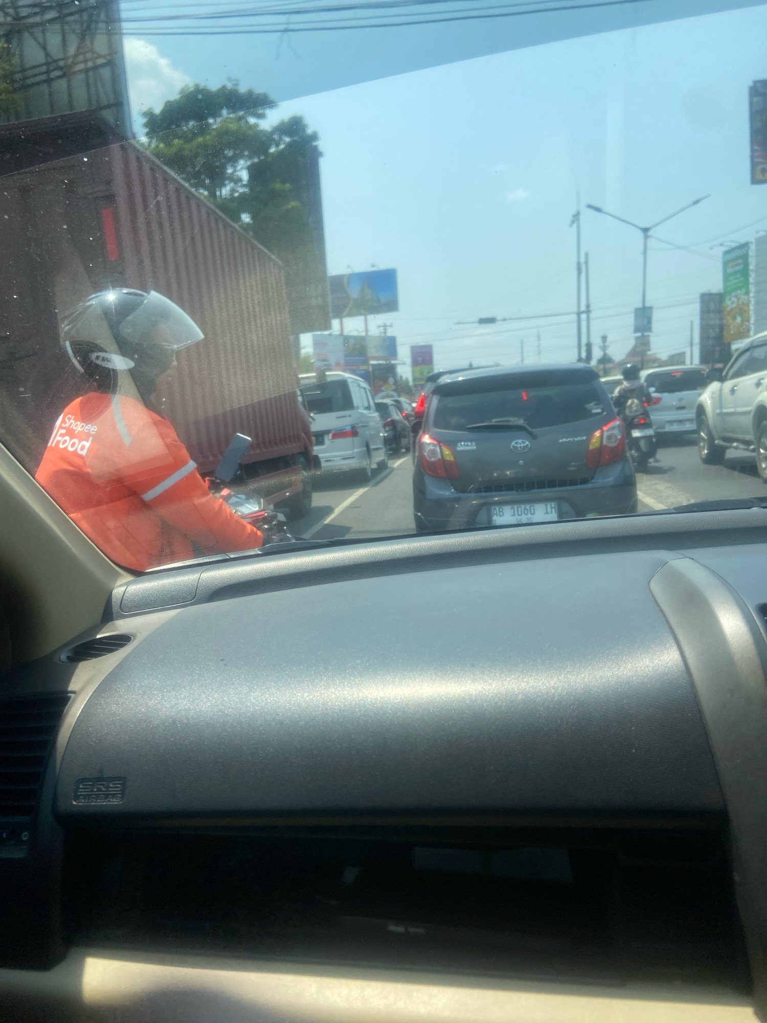 The image depicts a view from inside a vehicle, looking out through the windshield at a traffic jam on a sunny day. In the foreground, the dashboard of the car is visible, with an "SRS AIRBAG" label on the passenger side. Outside the vehicle, a motorcycle rider wearing an orange Shopee Food uniform and a helmet is in the left lane, next to a large red shipping container. Several other vehicles, including a dark gray car with license plate "AB 1060 IH", a white SUV, and other cars and motorcycles, are also present in the traffic jam. Buildings and trees are visible in the background, along with streetlights and a blue sky. The scene appears to be on a road in the city of Ngaglik, Indonesia.