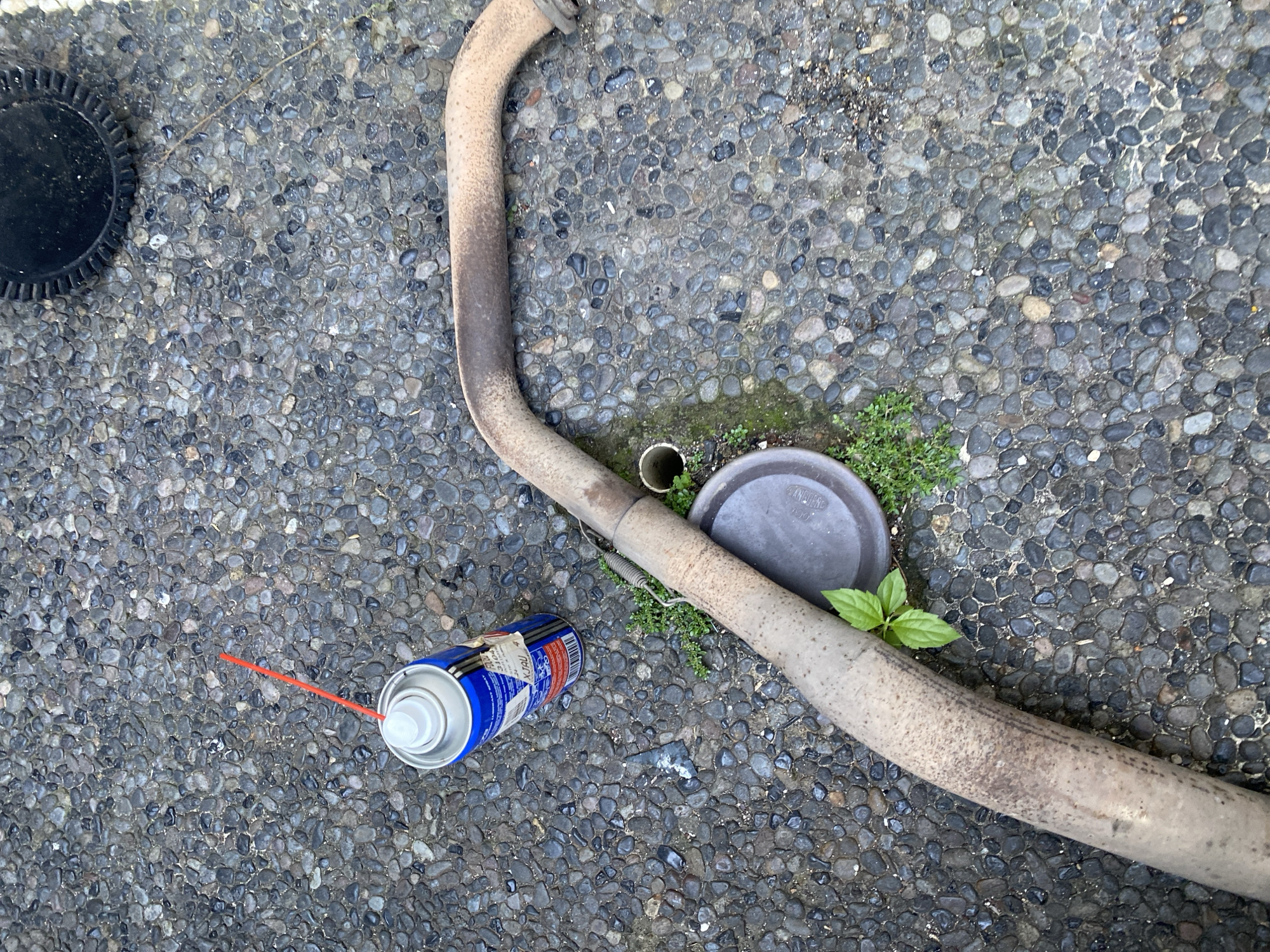 The image depicts an outdoor scene on a gravelly surface. A bent, rusty exhaust pipe lies on the ground, with a small green plant growing near its center. To the left of the exhaust pipe is a can of spray lubricant with its red nozzle extended. A dark circular object, possibly a lid or cap, is visible near the exhaust pipe and the spray can. The background is composed of the textured gravel. The location is stated to be Kuta Selatan, Indonesia.