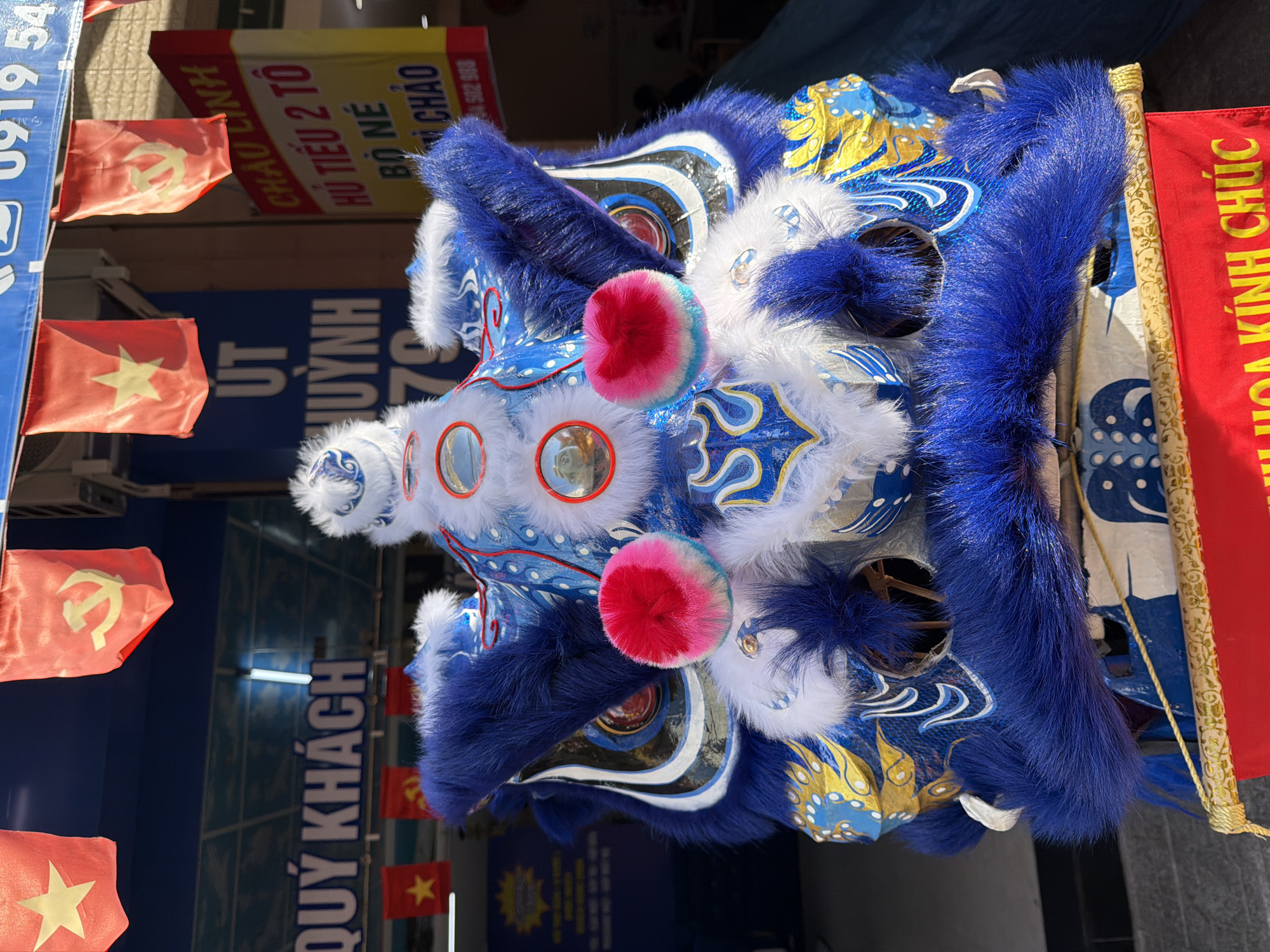 The image shows a close-up of a vibrantly colored lion dance costume, predominantly blue and white with gold accents. The costume is elaborate, featuring faux fur, reflective circular elements, and fluffy pink pom-poms. In the background, there are red flags with yellow stars and a hammer and sickle emblem, suggesting a cultural or festive context. Signs with Vietnamese text are visible, one with the words "QUÝ KHÁCH" and another featuring a phone number. The scene appears to be outdoors, possibly in a commercial area of Nha Trang Ward, Vietnam, given the visible signage. The lion costume is the primary subject, positioned centrally and filling most of the frame.