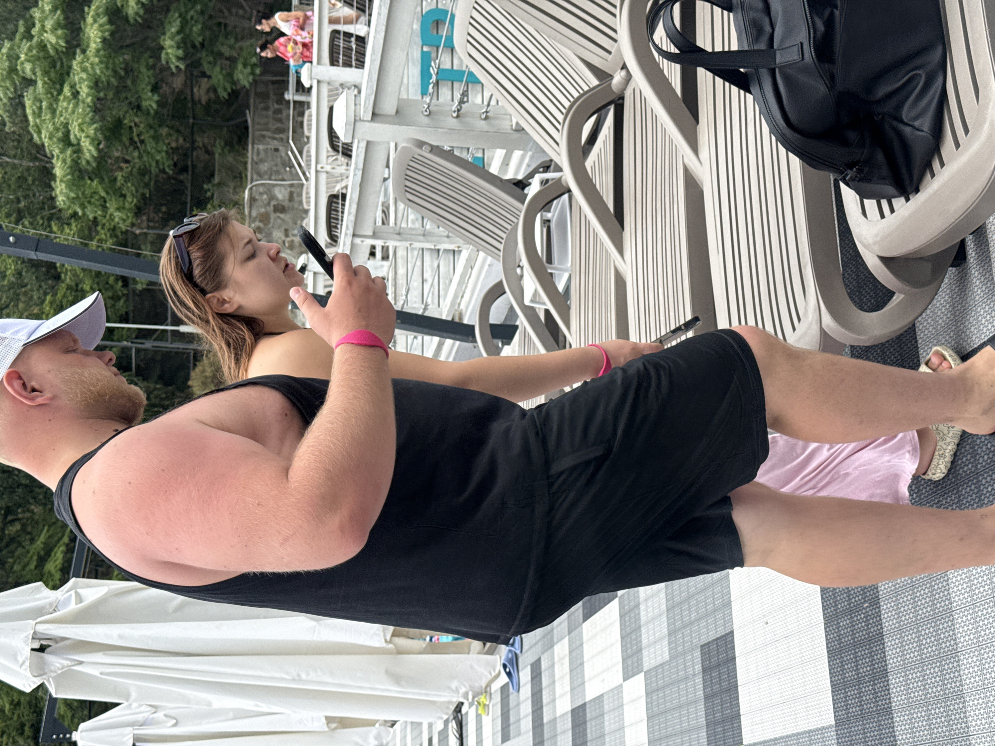 In Yalta, Ukraine, two individuals are standing on a patterned deck next to a white railing. The person on the left is a bald man wearing a black tank top, black shorts, and a white baseball cap. They are looking down at their phone. The person on the right is a woman with brown hair wearing sunglasses on her head. She is also looking down at a phone, possibly interacting with the man. A pink wristband is visible on the man's wrist. Behind them, several lounge chairs are set up, and in the background, there are trees and a body of water.