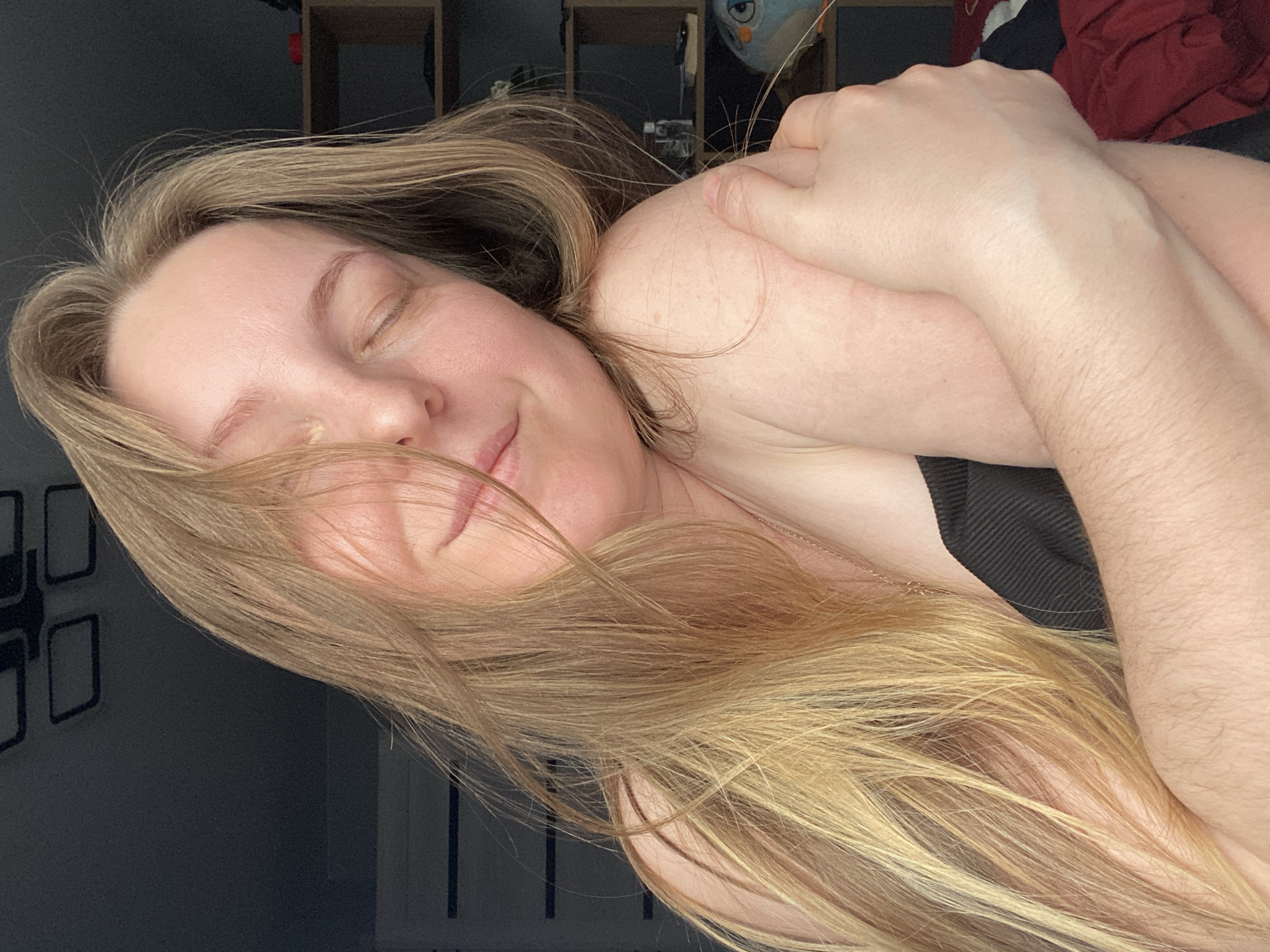 A close-up, angled portrait features a person with long, flowing light blonde to brown hair, with strands partially obscuring their face. Their eyes are closed, and a soft, gentle smile graces their lips, conveying a sense of serenity and contentment. The individual's arms are crossed, resting on their chest and shoulder, suggesting a comfortable or self-hugging posture. A delicate, gold-colored chain or necklace is visible against their skin, nestled amidst their hair. They appear to be wearing a dark, possibly black, top or garment strap.
The background is dark and softly lit, indicating an indoor setting in what appears to be a home in Noviy, Russia. On the upper right, a wooden shelving unit and a fluffy, owl-like plush toy with prominent eyes are partially visible. Below this, crumpled red and dark fabric can be seen. The left side of the background shows a dark wall with decorative square frames or outlines. The lighting is subdued and even, contributing to the tranquil atmosphere of the scene. No discernible text is visible within the image.