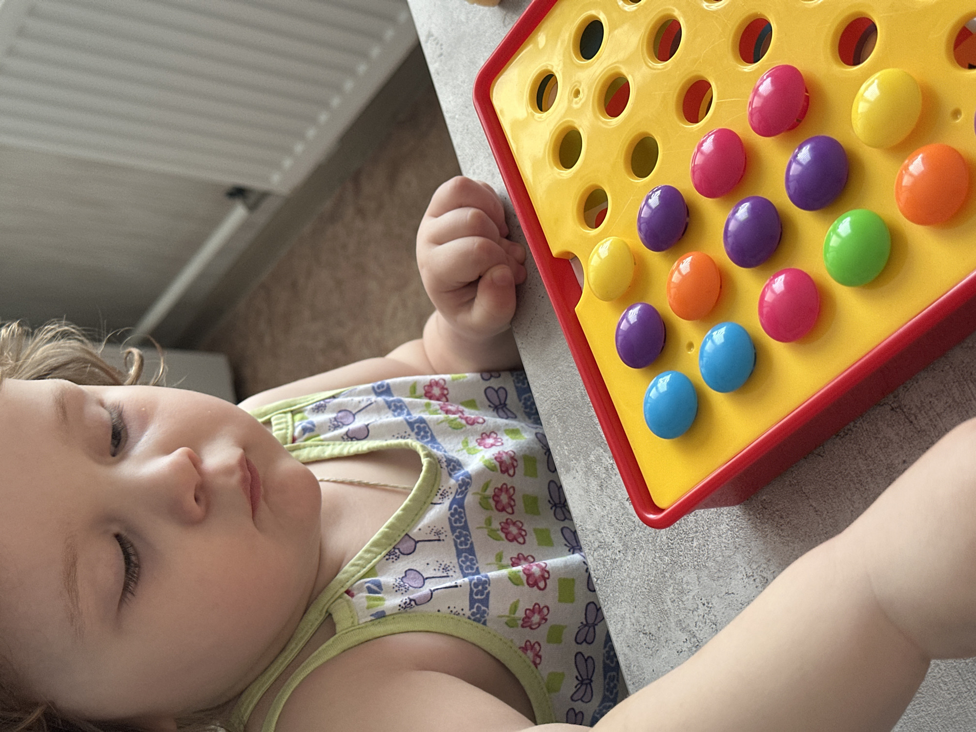 A baby is lying on a gray surface next to a yellow and red toy with colorful pegs in it. The baby is wearing a tank top with a floral and butterfly pattern. The location is possibly in Chaikovsky, Russia.